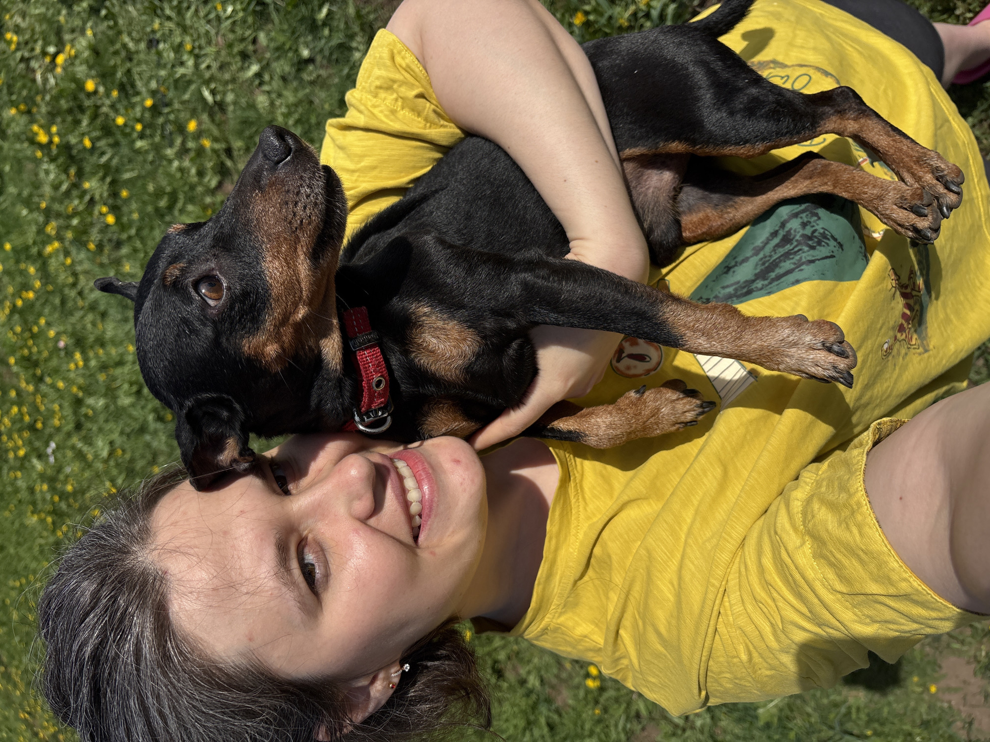 A woman is smiling up at the camera while holding a black and brown dog in her arms. The dog is wearing a red collar. The woman is wearing a yellow shirt and is laying in a grassy area with yellow flowers. The location is unknown and there is no indication of being in the city of Усть-Тунтор, Russia.