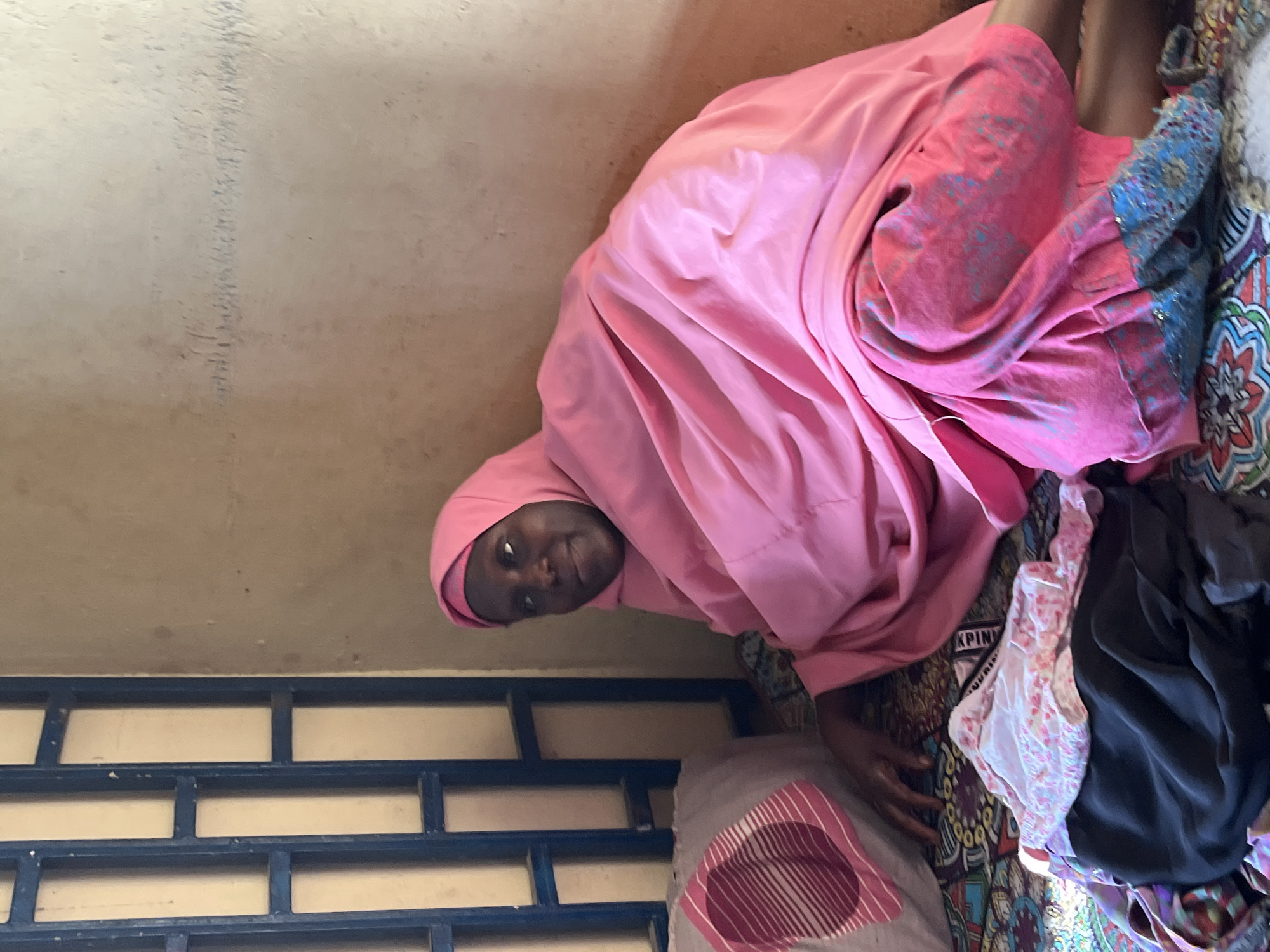 A woman wearing a pink headscarf and a matching pink garment is seated on a bed or mattress indoors. Her legs are visible, and she is partially obscured by various items of clothing, including a black garment, a floral patterned garment, and a gray pillow or cushion with a pink pattern. The background includes a beige wall with some markings and a section of a blue metal window or railing. The location is presumably in Jos, Nigeria.