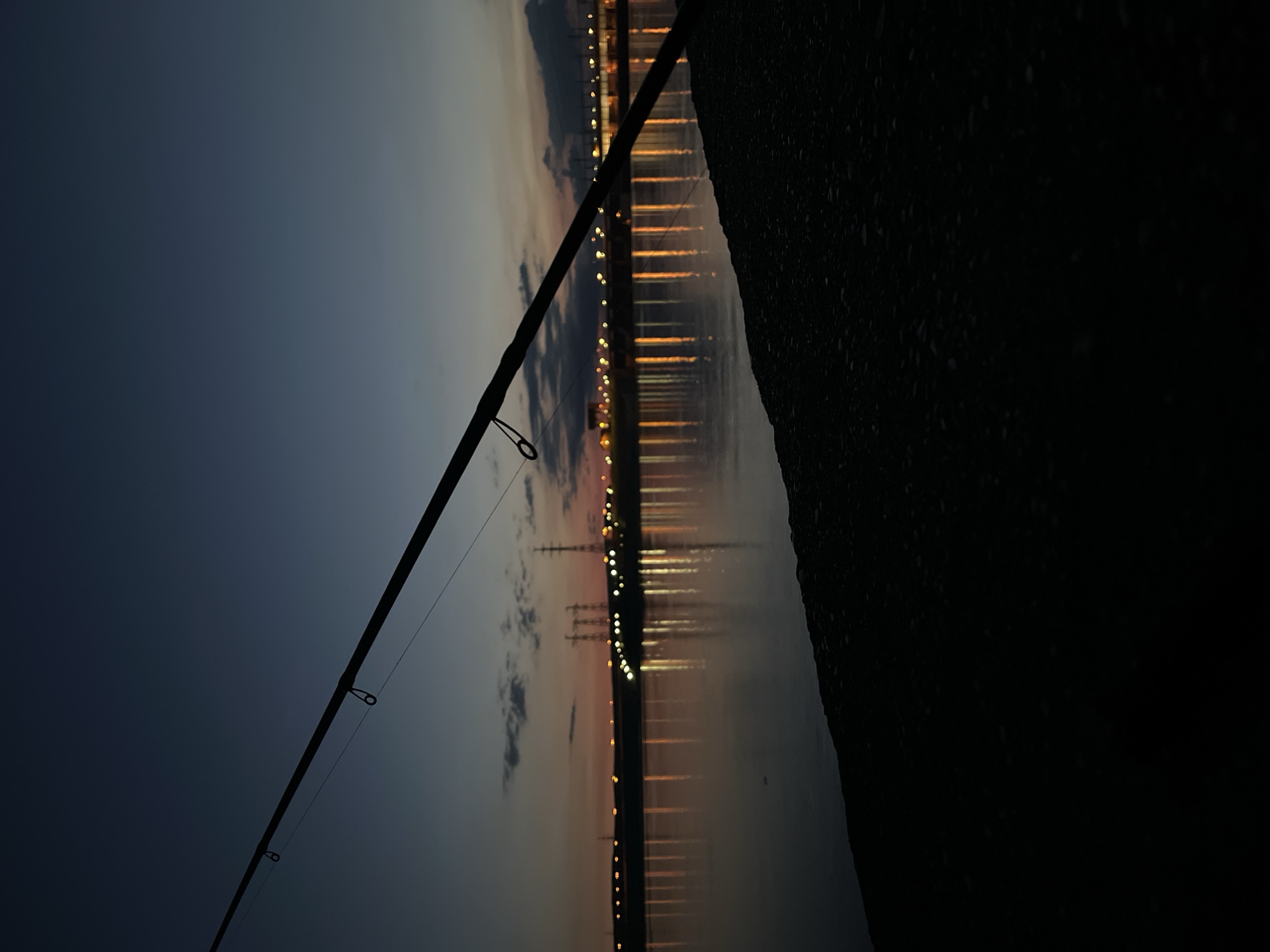 The image depicts a serene twilight or night scene by a large body of water, likely a river or reservoir, in Chaikovsky, Russia. A fishing rod is prominently silhouetted in the foreground, extending diagonally across the frame from the lower left. Its line guides are discernible, suggesting fishing is either in progress or recently concluded.
The sky exhibits a gradient of colors, transitioning from a deep, dusky blue-grey at the top to warmer orange and yellow hues near the horizon, indicative of either late dusk or very early dawn. Patches of clouds are visible against the fading light.
Across the calm water, the distant bank is lined with numerous artificial lights, possibly from a town, industrial complex, or a bridge. These lights cast elongated, shimmering reflections vertically across the water's surface, highlighting its stillness. Several electricity pylons or transmission towers are silhouetted against the illuminated horizon on the far bank. The immediate foreground on the right side of the image is a dark, textured area, likely the shoreline or bank where the photographer is situated. The scene evokes a peaceful and solitary atmosphere. No people or text are visible.