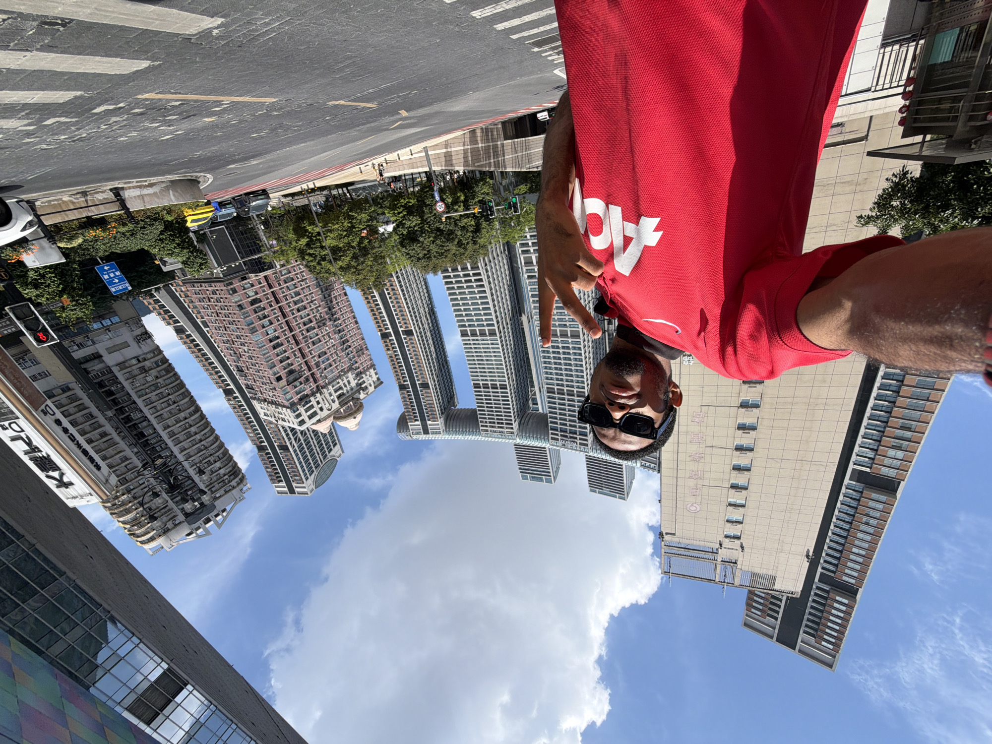 In Yuzhong District, China, a man wearing sunglasses and a red t-shirt with a white logo is taking a selfie. He is standing on a street with a road and crosswalk in the foreground. Behind him, several tall, modern buildings with distinct architectural designs dominate the skyline. The sky is blue with scattered white clouds. In the background, there are trees, traffic lights, and cars. The man is holding up two fingers in a peace sign.