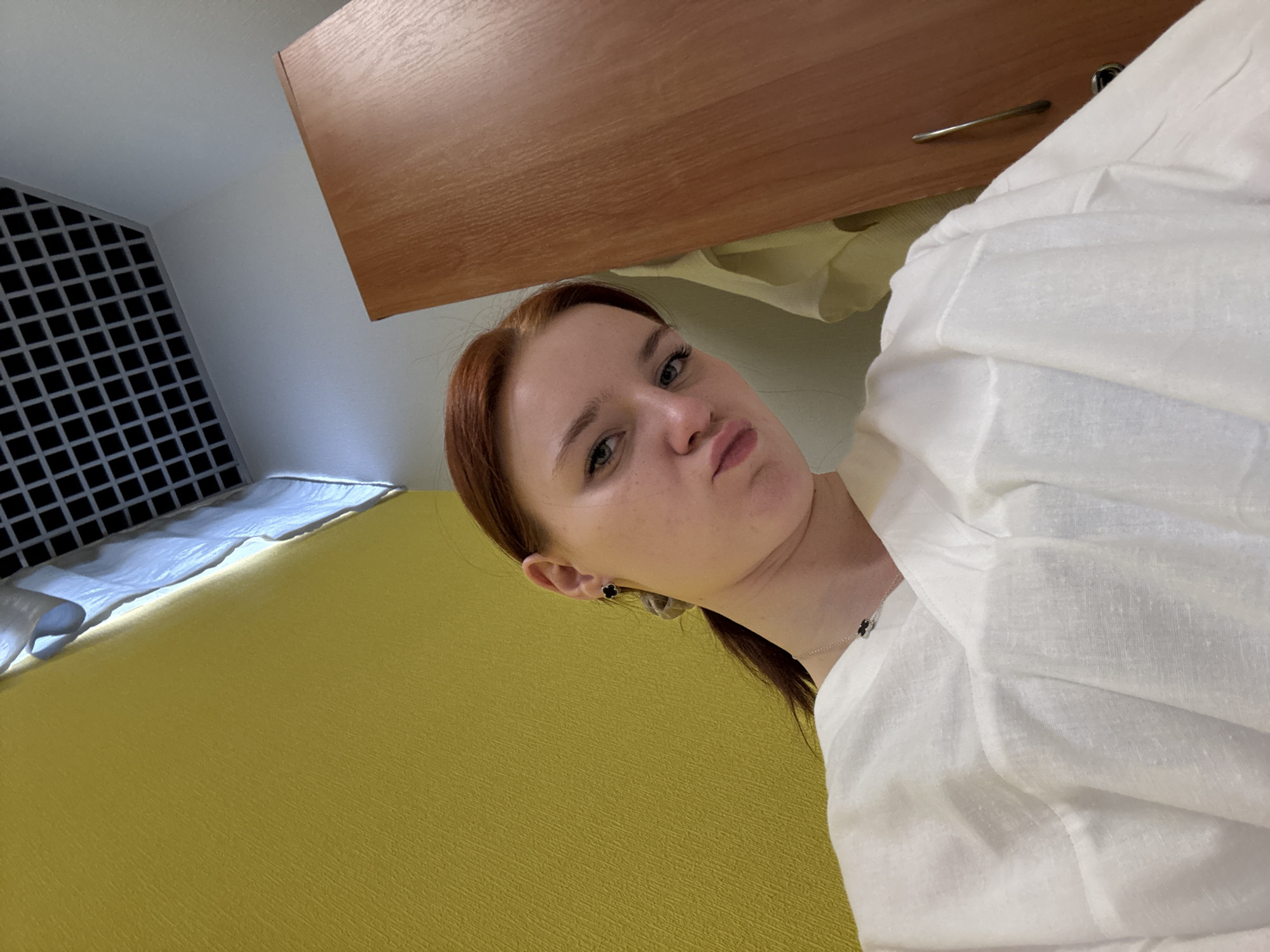 The image shows a young woman indoors, likely in a hotel room. She has reddish-brown hair, fair skin, and is wearing what appears to be a white linen shirt or dress. She has a neutral expression, with her lips pursed slightly. She is wearing earrings and a necklace with a small clover or flower pendant.
The background reveals parts of a room:
- a yellow-colored bed or surface.
- a black and white ventilation or cooling system on the wall.
- a wooden cabinet or drawer.
- white sheets or covers are partly visible on the bed.
The photo's angle is unusual, suggesting it might be a selfie taken at an odd tilt. The lack of distinct features prevents a concrete location within Saint Petersburg, Russia, although it might be possible to determine the specific hotel using more details.