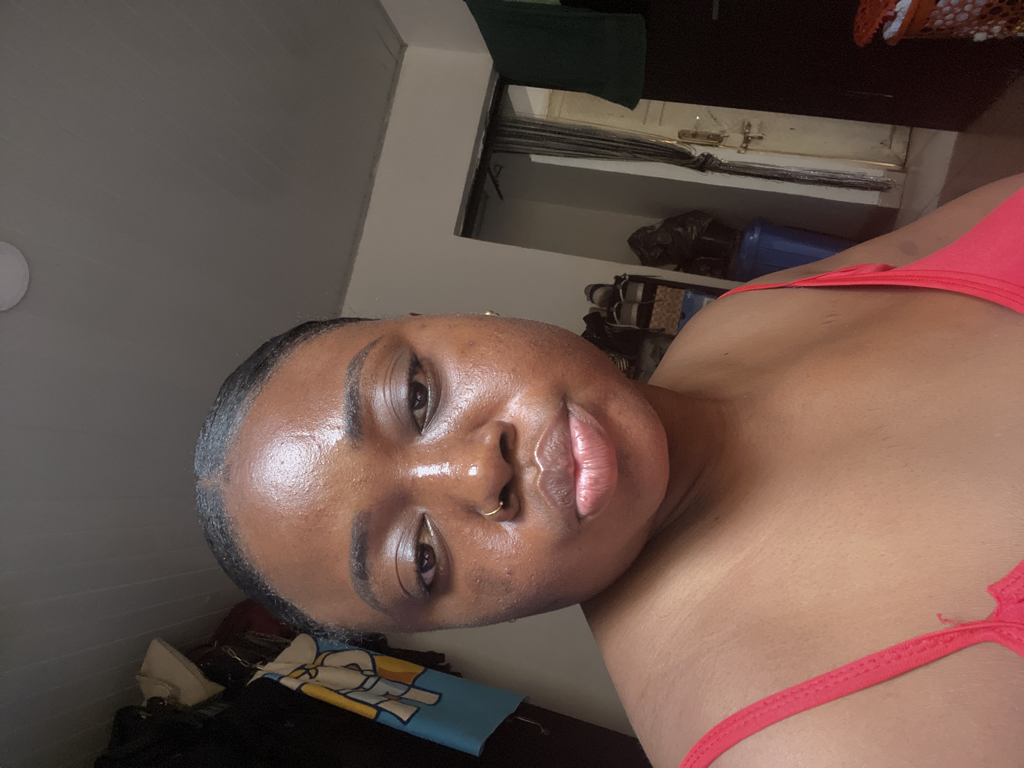 A close-up, angled shot depicts a woman in a red top. Her face is front and center, showing a gold nose ring and slightly glossy skin. The background includes a bedroom setting with a closet and a cluttered space. There is no evidence in the image to indicate the location as Abuja, Nigeria.