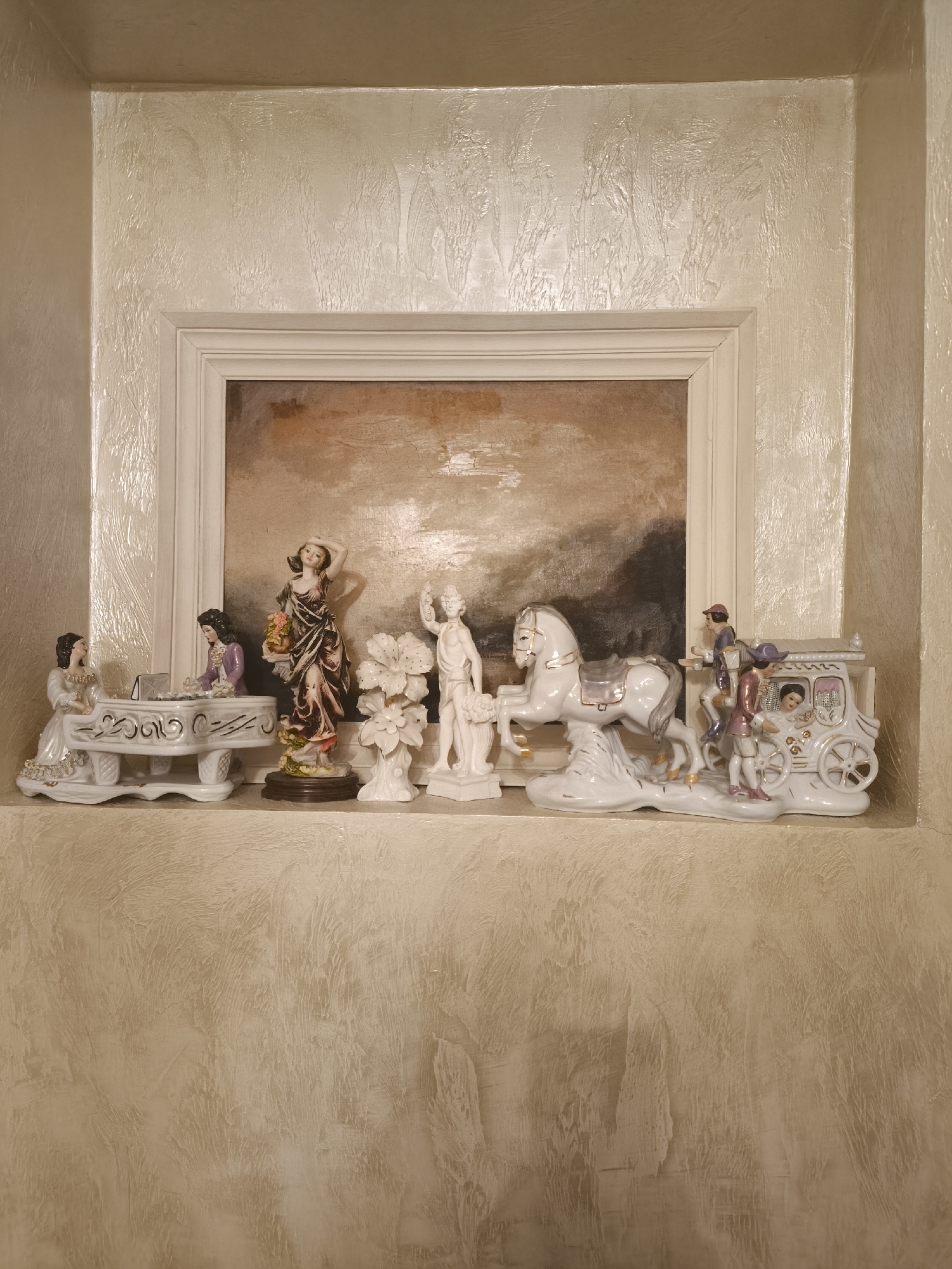 A decorative alcove in a building in Batumi, Georgia, features a framed painting with an abstract, earthy-toned landscape. A shelf below the painting displays several porcelain figurines. From left to right, the figurines depict: two women seated at a white piano with ornate gold detailing; a woman in a patterned dress holding a bouquet of flowers; a statue of a man holding flowers; a white horse and carriage with two figures inside and one figure standing beside it. The wall surrounding the alcove is textured with a stucco finish in a light, neutral tone.