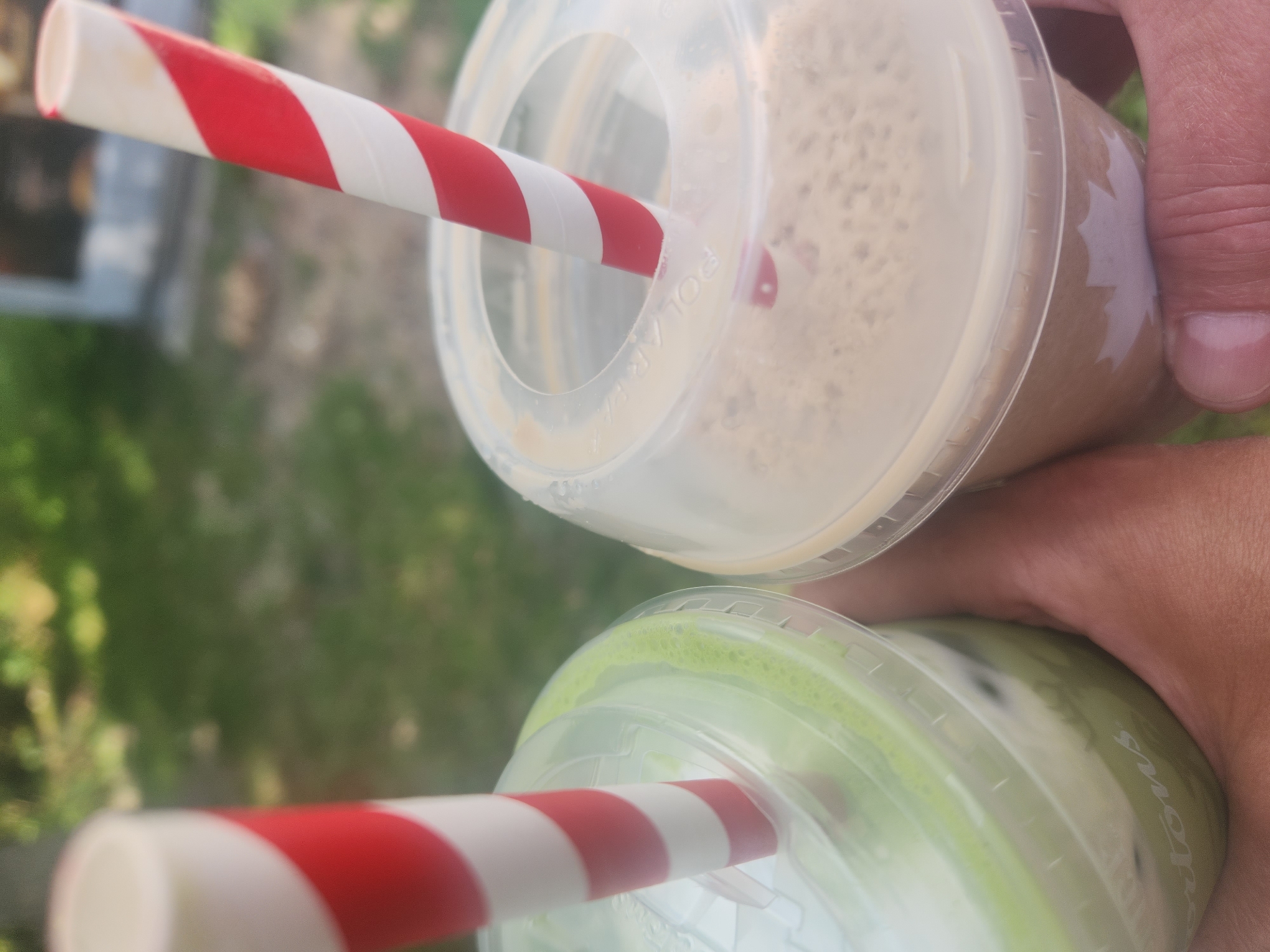 The image shows two iced drinks with red and white striped straws held by hands. The drink on the left is green and appears to be a matcha latte. The drink on the right is a light brown color and could be an iced coffee or frappuccino. Both drinks are in clear plastic cups with plastic lids. The background is blurred and appears to be an outdoor setting with green foliage, suggesting a park or a patio. There is text on the cup on the left that appears to say "Tims," likely indicating Tim Hortons, a popular Canadian coffee chain. The image does not provide any specific landmarks or visual cues to pinpoint a precise location within the city of Toronto, Canada, but the presence of Tim Hortons strongly suggests it is in Canada.