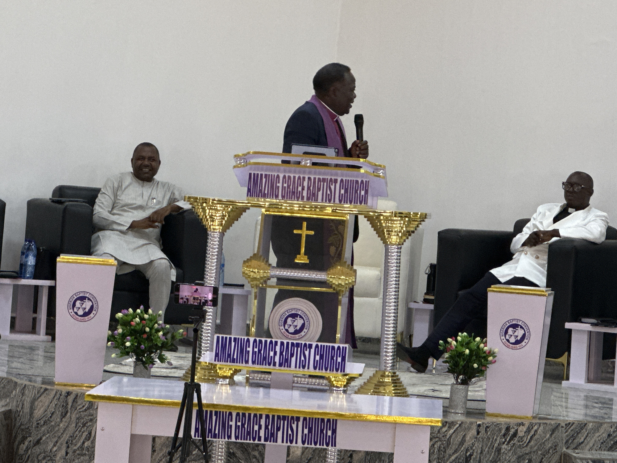 The image depicts an indoor religious gathering at the Amazing Grace Baptist Church in Goni Gara, Nigeria. The scene is set on an elevated stage with a dark, mottled surface against a plain white wall background.
In the central-right portion of the frame, a dark-complexioned man, appearing middle-aged to senior, stands behind an elaborate white and gold multi-tiered podium. He is dressed in a dark suit with a purple stole draped over his shoulders and is actively speaking into a black handheld microphone, with his mouth open. The podium prominently displays the text "AMAZING GRACE BAPTIST CHURCH" on its front, features a gold cross on a dark background in its central section, and behind it, a circular emblem indicating "NIGERIAN BAPTIST CONVENTION ESTABLISHED 1914."
To the speaker's left, a dark-complexioned man, appearing middle-aged, is seated in a black armchair. He wears a light-colored traditional garment and is smiling, with his arms resting on the chair's armrests. To the speaker's right, another dark-complexioned man, also appearing middle-aged to senior, is seated in a black armchair. He wears a white clerical shirt with a black collar, dark trousers, and dark-framed glasses, with his hands resting in his lap.
Small white tables with gold trim are positioned on either side of the stage. A blue plastic water bottle is visible on the table to the left. On the lower stage level, two small arrangements of artificial white, pink, and purple flowers in metallic pots flank the podium. A smartphone mounted on a black tripod is positioned on the far left of the stage, recording the event.