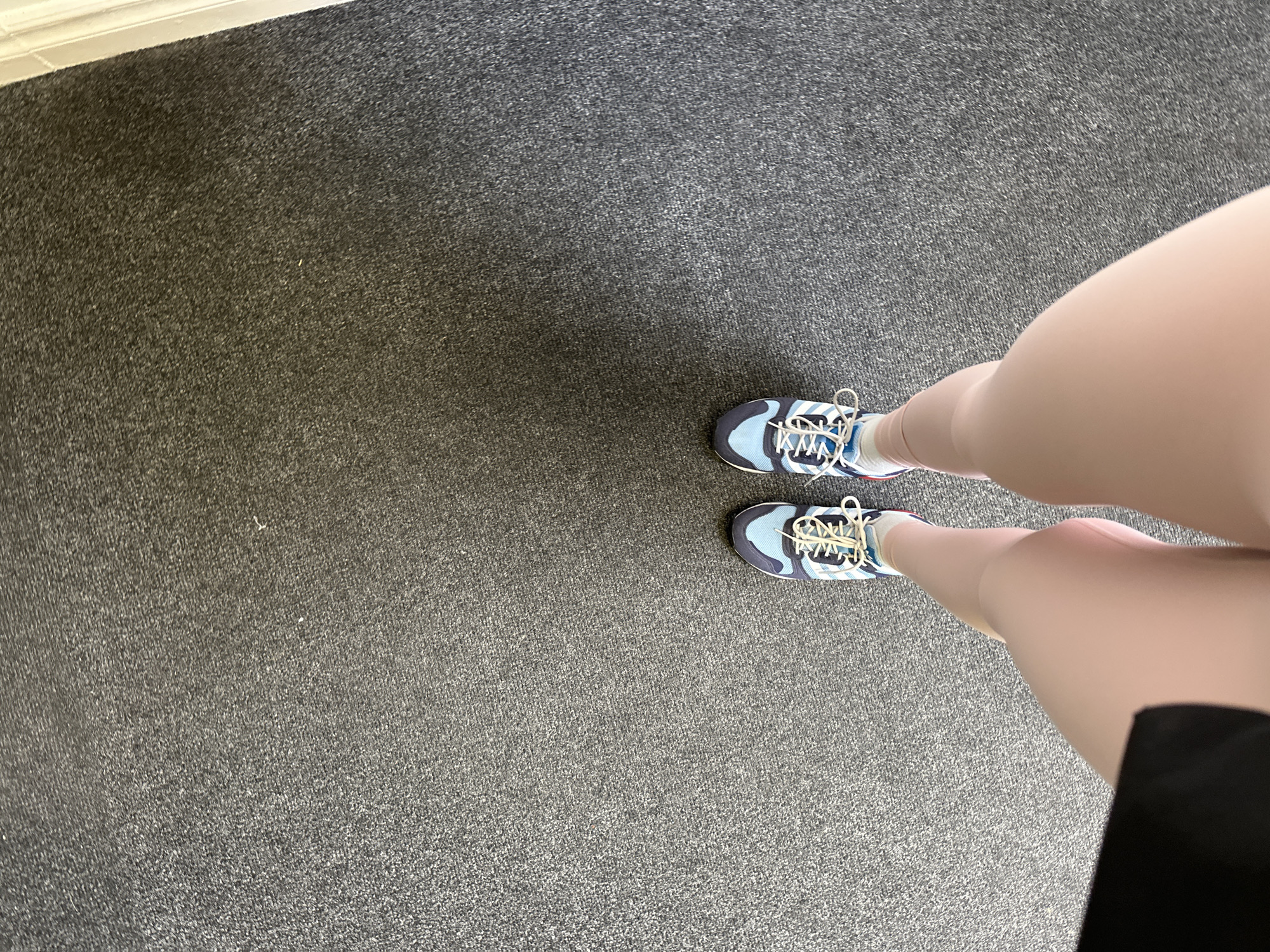The image shows a person's legs and feet, wearing blue and white athletic shoes, standing on a gray carpeted floor. The perspective is from above, looking down at the person's feet. The scene is indoors, with a white trim visible on the right side of the image, possibly indicating a doorway or wall. The lighting suggests a well-lit indoor environment. No specific landmarks or details are visible to identify a location within the city of Carlow, Ireland.
