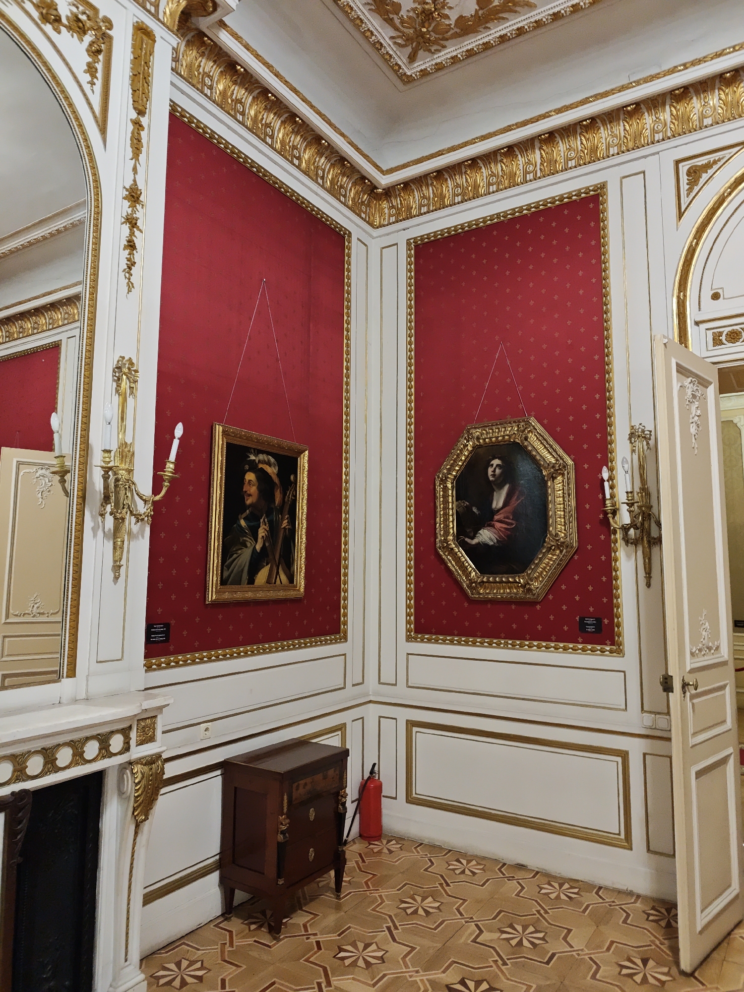 The image depicts an opulent interior space, likely a museum or a historic residence, characterized by rich red walls adorned with a repeating pattern, and ornate gold detailing on the walls, ceiling, and trim. Two framed paintings are prominently displayed on the red walls: one a rectangular portrait of a man with a mustache and feathered hat, holding a musical instrument, and the other an octagonal portrait of a woman looking upwards. A gilded sconce with two electric candles is mounted on the wall near the rectangular painting, and another similar sconce is visible near the octagonal painting. To the left, a large mirror framed in gold reflects the room. A white fireplace with gilded decorative elements is on the far left. In the foreground, a dark wooden chest of drawers with brass accents stands on a patterned wooden floor. A red fire extinguisher is visible near the chest of drawers. An open door with a cream-colored frame and decorative molding is on the right side of the image. The ceiling features intricate gilded stucco work. The overall impression is one of grandeur and historical significance. This scene is likely located within the Lviv National Art Gallery, specifically in one of its historic exhibition halls in Lviv, Ukraine, which houses a significant collection of European art.