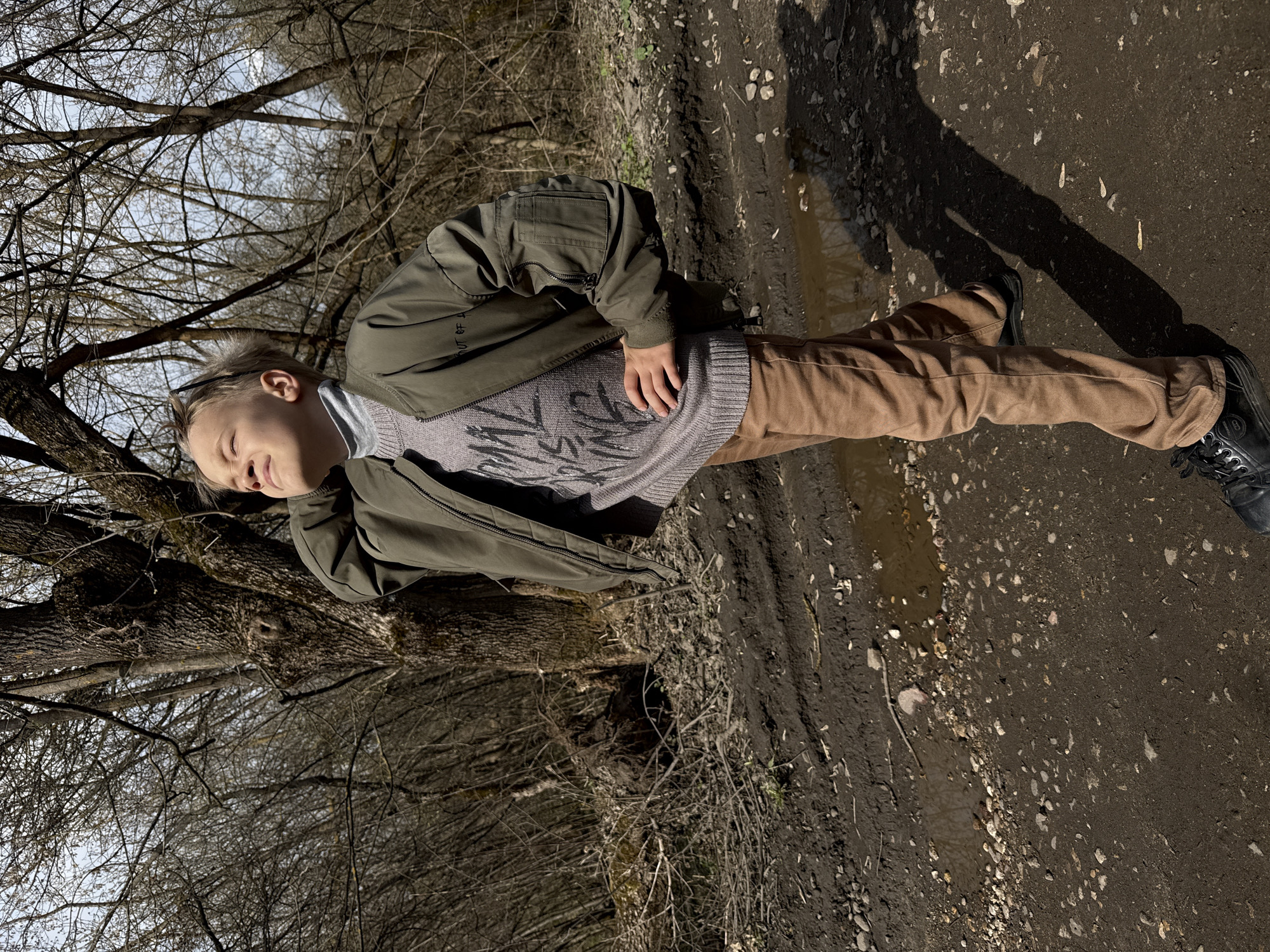 A young person stands in a natural setting, appearing to be near a wooded area. The individual is dressed in a khaki-colored jacket over a gray sweater and brown pants, with dark-colored shoes. The ground consists of mud and dirt, with some puddles. The trees are bare, suggesting it is either late autumn or early spring. The location appears to be in a park-like area. The location in Dmitrov, Russia, cannot be verified without more information from the image.