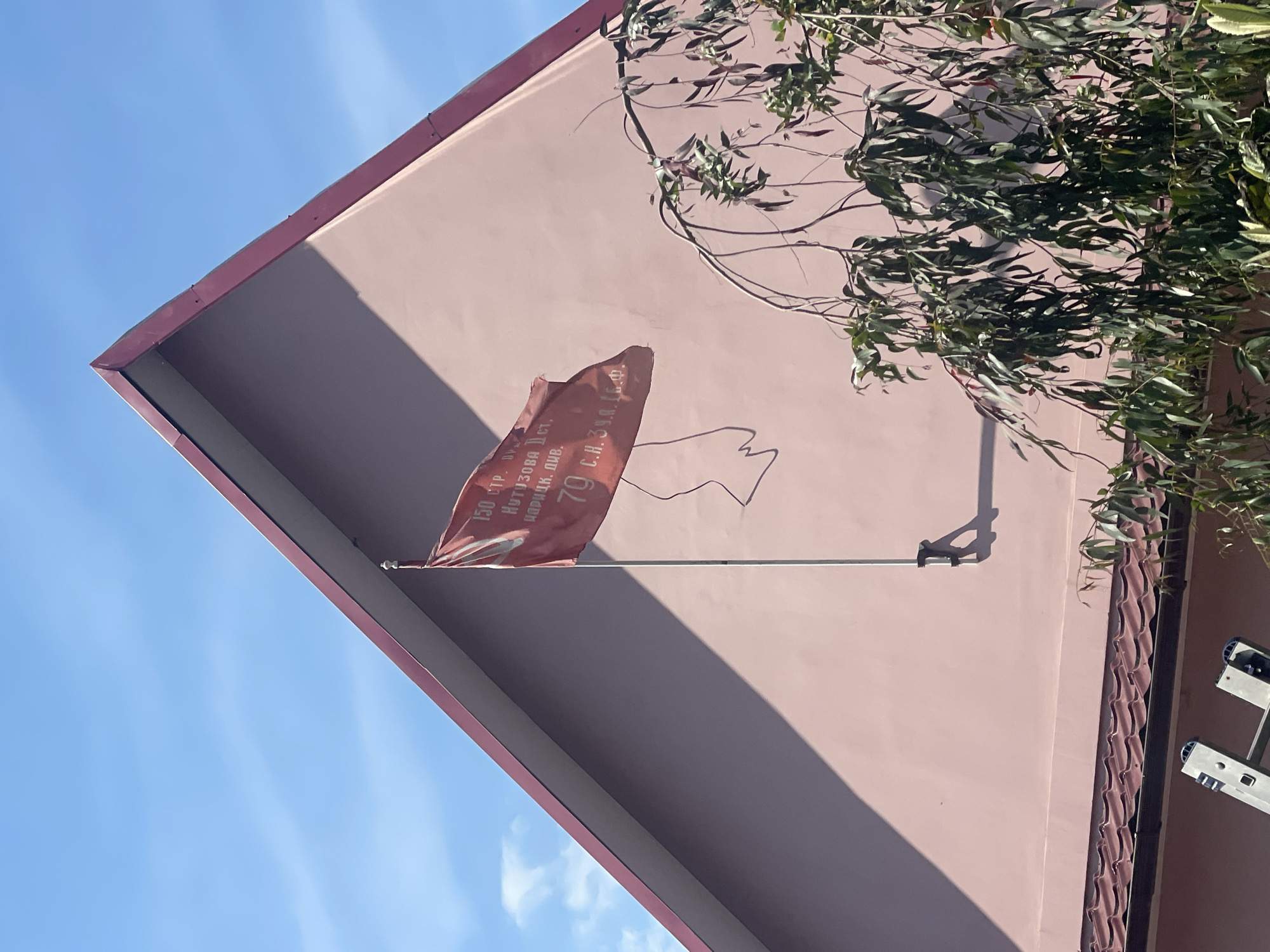 The image depicts the gable-end wall of a building in Sochi, Russia, under a clear blue sky with sparse, high-altitude clouds. The wall is painted a light reddish-brown or dusty rose color, with a prominent triangular shadow cast by the roof overhang on its left side, indicating direct sunlight from the upper left. The roofline is trimmed with a dark reddish-brown material.
A reddish-orange flag, appearing somewhat worn or tattered, is mounted vertically on a pole attached to the wall. White Cyrillic text is visible on the flag, partially obscured by folds. The discernible text reads: "150 СТР." (150th Rifle), "КУТУЗОВА I СТ." (Kutuzov 1st Class), and "ИДРИЦКАЯ. ДИВ." (Idritskaya. Division.), which refers to the 150th Idritsa Rifle Division, a decorated unit of the Great Patriotic War. Below this, a larger number "79" is visible, followed by "С.Н.З. И.В. 1.Ф." This likely commemorates the 79th anniversary of Victory in World War II (Great Patriotic War), with the subsequent letters potentially denoting further military honors or a specific unit sub-designation.
In the lower right, branches from a tree with long, dark green, pendulous leaves extend into the frame. The top section of a silver metallic extendable ladder is partially visible in the bottom-left corner of the image, suggesting recent or ongoing work on the building. The overall scene conveys a commemorative or patriotic display on a building, likely a private residence or a small public structure, during daytime with good weather.