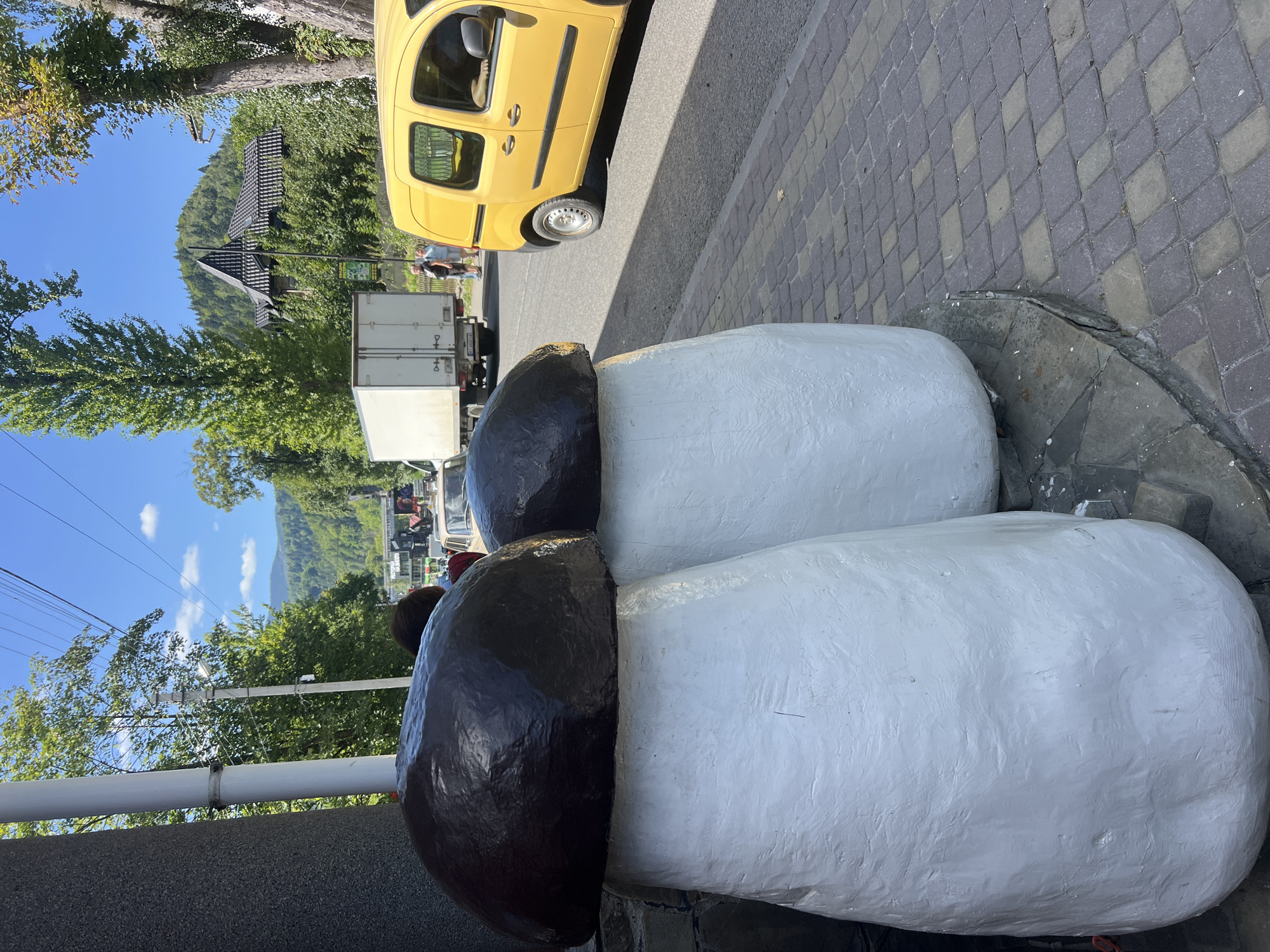 The image depicts a street scene in Yaremche, Ukraine, featuring two oversized mushroom sculptures as the foreground subjects. These sculptures, painted white with dark brown caps, are situated on a paved walkway. Behind them, a street runs horizontally, with a yellow van and a white truck parked on it. Several people are visible near the vehicles. The background reveals a densely forested hillside with a traditional wooden house with a dark roof. The sky is blue with scattered clouds, and overhead power lines are present. The overall environment suggests a sunny day in a town nestled among mountains.