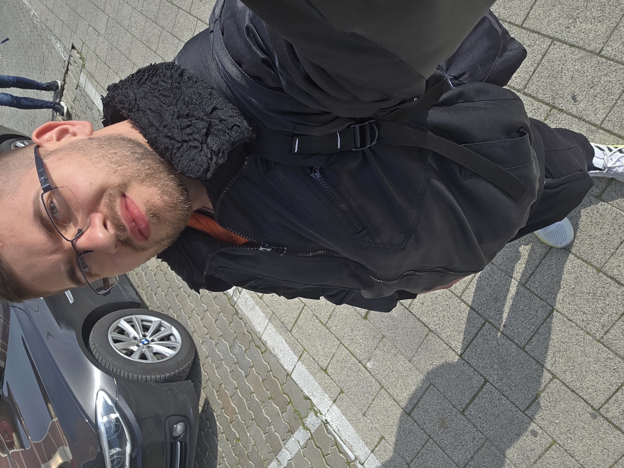 A man with short brown hair and glasses stands next to a dark-colored car, with a portion of the front wheel visible. He is wearing a black jacket with a fur-lined collar and black pants. He is looking into the camera. The background shows a paved surface with white painted lines, and in the distance, two people are walking. The location appears to be outdoors. Specific identification of the location as Plovdiv, Bulgaria, is not verifiable from the image alone.