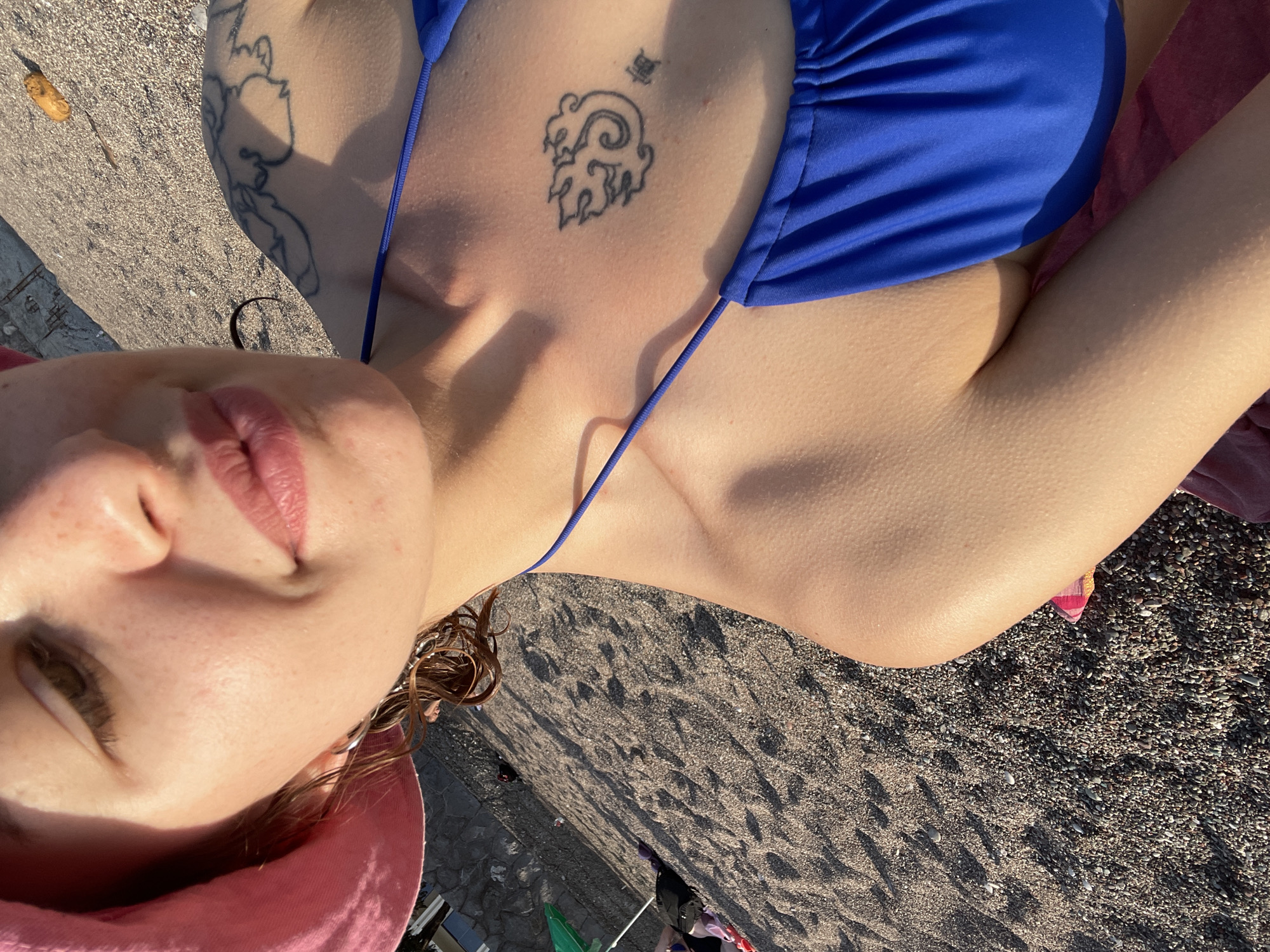 A woman wearing a blue bikini and a pink hat is lying on a beach in Rafailovići, Montenegro. She has a tattoo on her chest and another on her arm. The beach is made up of small pebbles. The sun is shining, casting shadows on the beach. There are other people and objects in the background, including an umbrella and some bags.