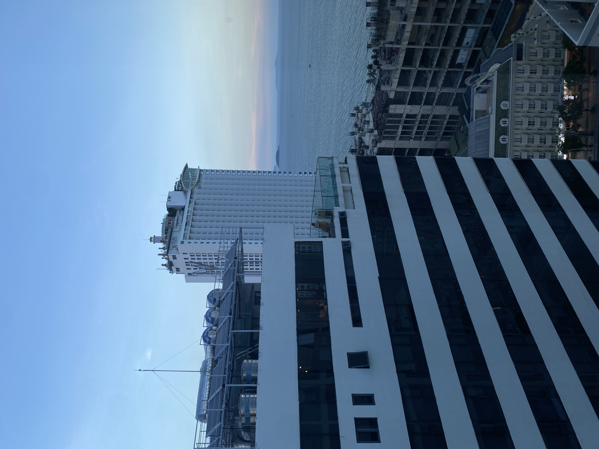 The image shows a city skyline with a body of water in the background, likely the sea. In the foreground, there is a building with a distinctive black and white striped facade. Above and behind this building, a taller, modern white building with many windows is visible. The top of this building appears to be a hotel or resort, with visible outdoor seating and structures. Further in the background, there are unfinished or under-construction buildings, suggesting ongoing development in the area. The sea is calm, with a few small boats visible on the horizon. The sky is a dusky blue, indicating either dawn or dusk. The overall scene depicts a coastal urban environment. The text on the water tanks on the roof of the foreground building appears to say "HOANG HAI," which translates to "Yellow Sea" in Vietnamese, potentially indicating a local business or product. The presence of multiple tall buildings and the proximity to the sea suggests a location like Nha Trang Ward, Vietnam, a popular coastal resort city known for its beaches and modern architecture.