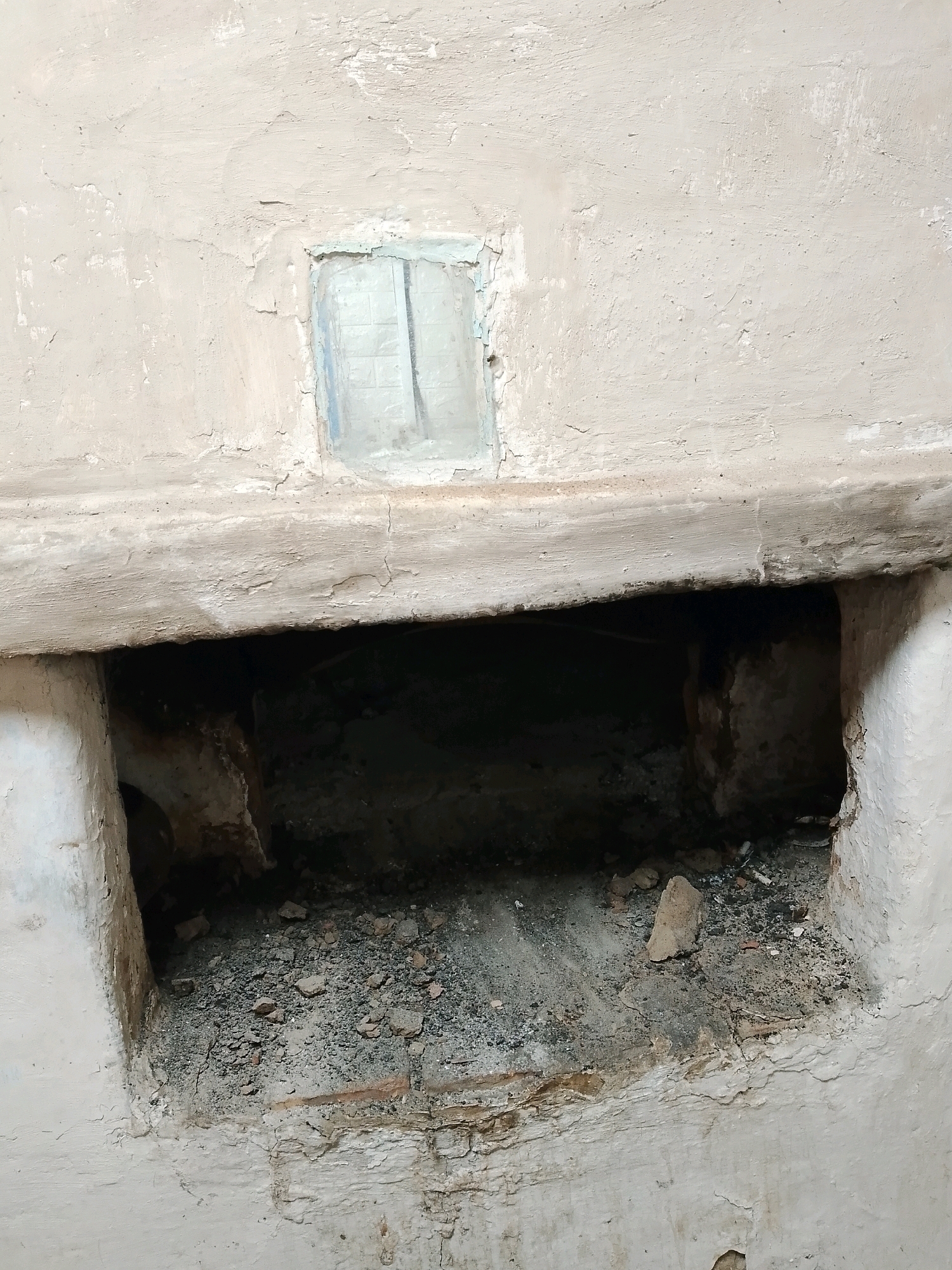 A close-up, eye-level shot shows a partially damaged, light-colored wall with a large, dark, rectangular opening at the bottom. The opening appears to be a void or cavity within the wall, filled with debris. Above the opening, there is a thick, rough-textured ledge that spans the width of the frame. In the upper center of the wall, a small, rectangular pane of glass is set into a recessed area. The wall's surface is textured and shows signs of wear, with peeling paint and cracks. The overall impression is one of disrepair and abandonment. The image was taken in the vicinity of Ленінскі сельскі Савет, Belarus.