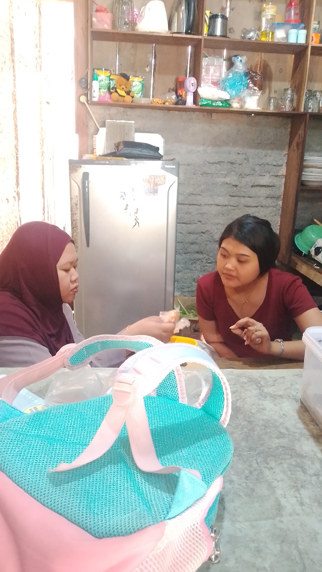 In Karanglangu, Indonesia, two women are seated at a table in a rustic kitchen. The woman on the left, wearing a maroon hijab and gray top, is looking down and holding food. The woman on the right, wearing a maroon v-neck shirt, is looking towards the left and gesturing with her right hand. In the foreground, a teal and pink mesh bag with straps is placed on the table. Behind the women, a silver refrigerator stands against a brick wall. Shelves above the refrigerator are filled with various items, including a teddy bear, food packages, bottles, and kitchenware. The environment suggests a casual, domestic setting within a home.
