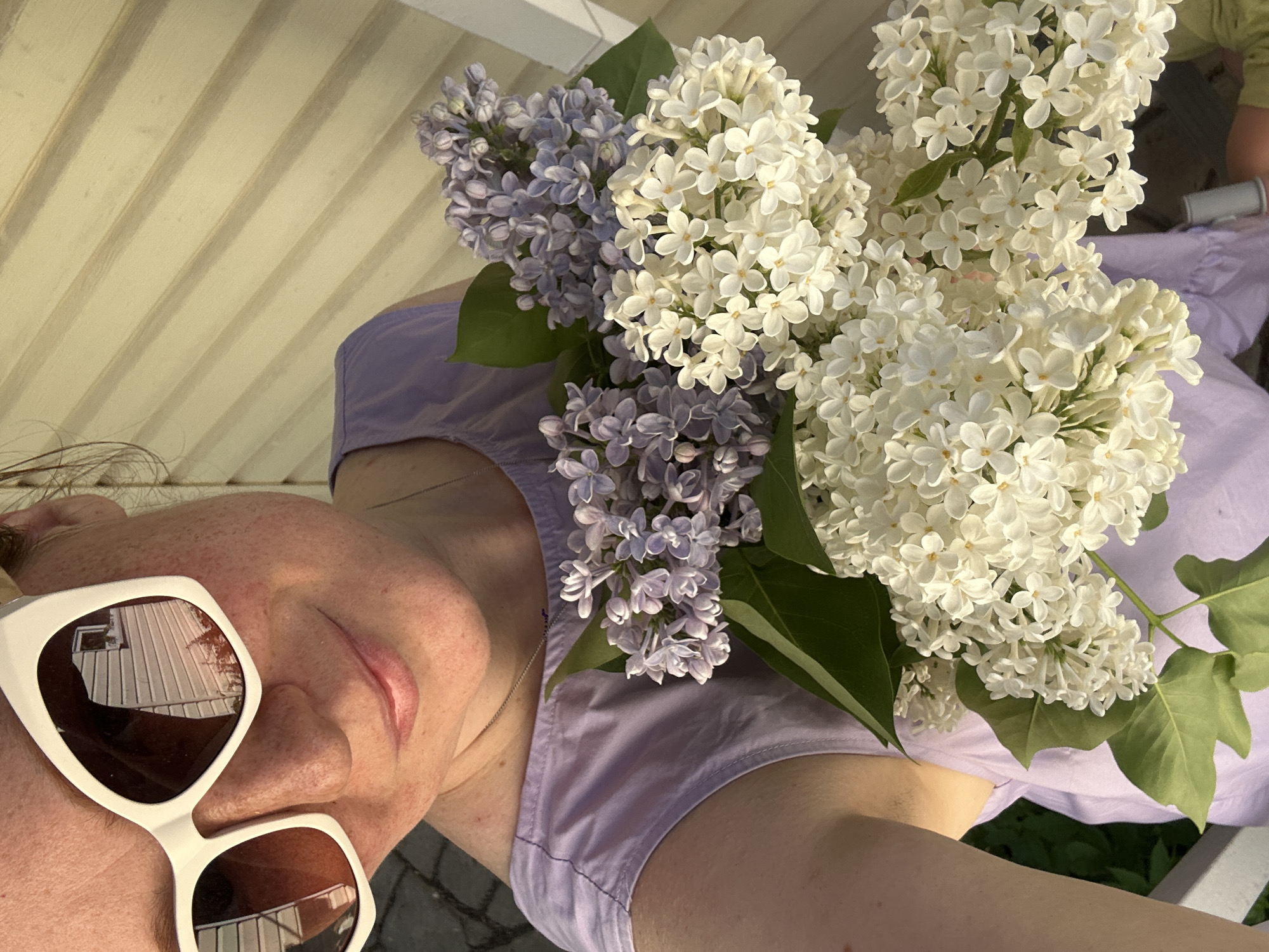 A selfie features a person wearing white sunglasses and a lilac-colored dress, holding a bouquet of purple and white lilacs. The reflection in the sunglasses shows a building with white siding and window frames. The background includes a section of wooden siding similar in color to the building's reflected siding. Another person is visible in the background.