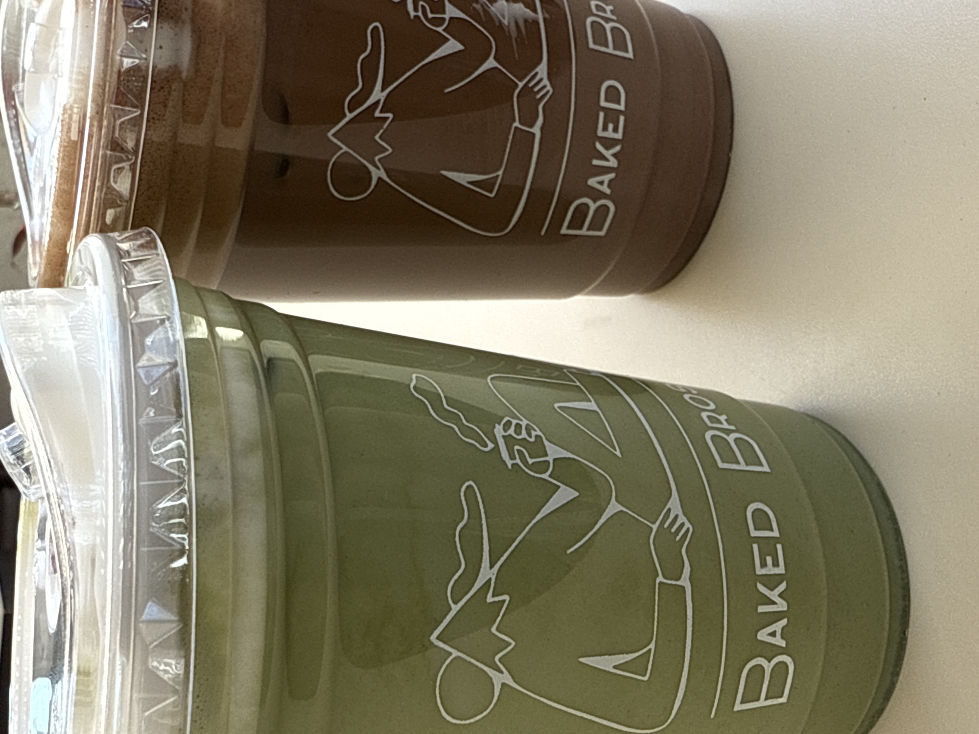 Two plastic cups containing beverages are positioned on a light-colored surface in San Bernardino Tlaxcalancingo, Mexico. The foreground cup on the left is olive green, filled with a light green liquid topped by a creamy white layer, and sealed with a clear plastic dome lid. It features a white line-art illustration of a stylized figure with a mountain head, seated and holding a cup from which vapor rises, with the text "BAKED BRO" printed below. Partially visible to the right and slightly behind is a dark brown plastic cup, also with a clear plastic dome lid, containing a dark brown liquid with a frothy top layer. This cup displays a similar white line-art illustration and partial "BAKED BRO" text. The surface appears flat and uniform.