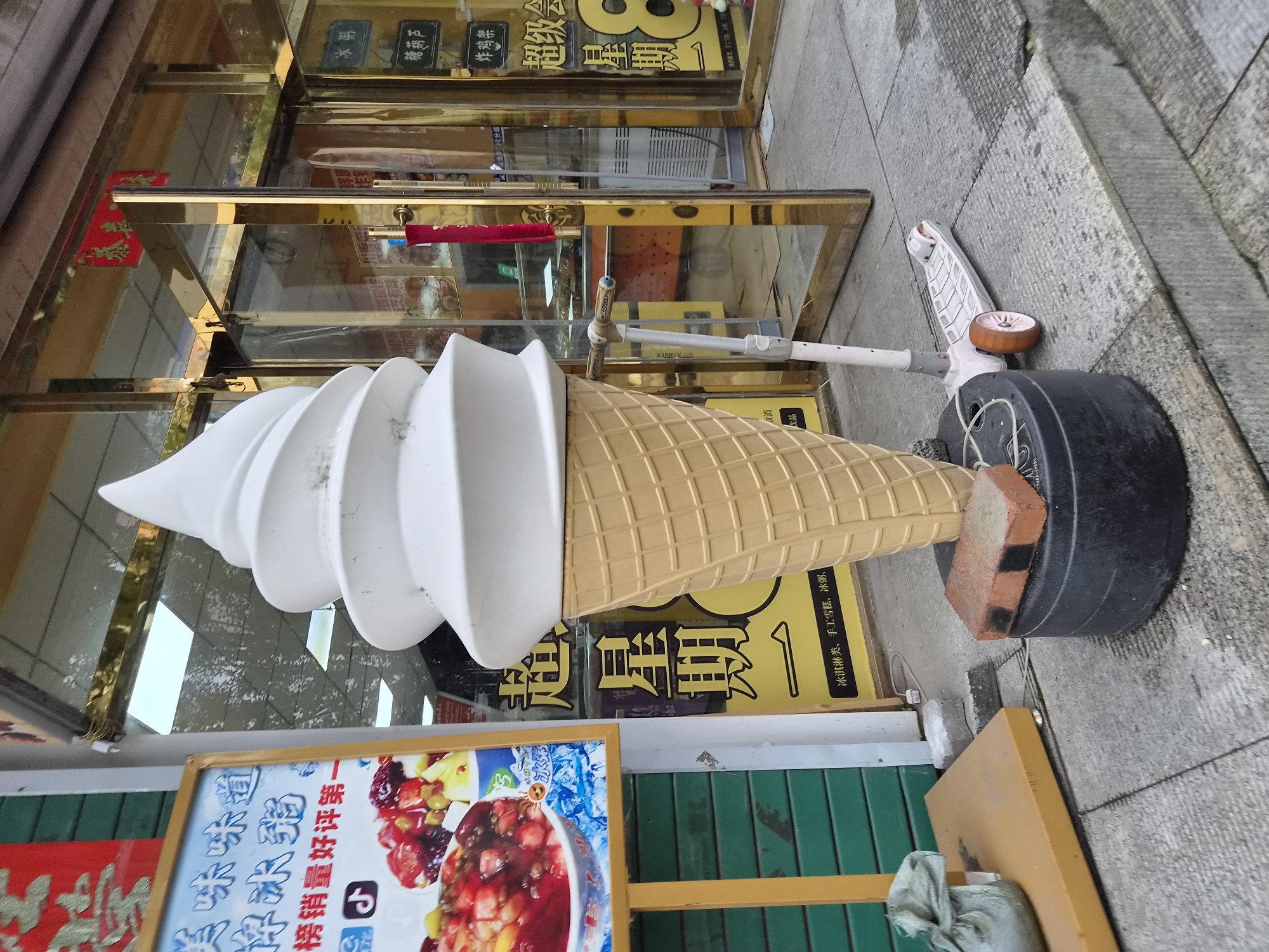 A large, white soft-serve ice cream cone sculpture is the focal point of this outdoor scene in front of a shop in Hunchun, China. The ice cream cone is on a black circular base, with a brick and a part of a scooter resting on or near it. A white kick scooter with pink accents is positioned to the right of the ice cream cone. To the left of the ice cream cone, a yellow sign with Chinese text and an image of a fruit salad is displayed. Behind the sign, a green corrugated metal shutter is visible. The shop entrance, with a gold-colored frame, is to the right of the ice cream cone, and through the glass doors, a display area with shelves and products can be seen. Several signs with Chinese text are posted on the shop's facade. The ground is paved with gray square tiles and concrete. A gray fabric bag is placed on a yellow platform to the left of the ice cream cone. The overall environment appears to be a commercial street.