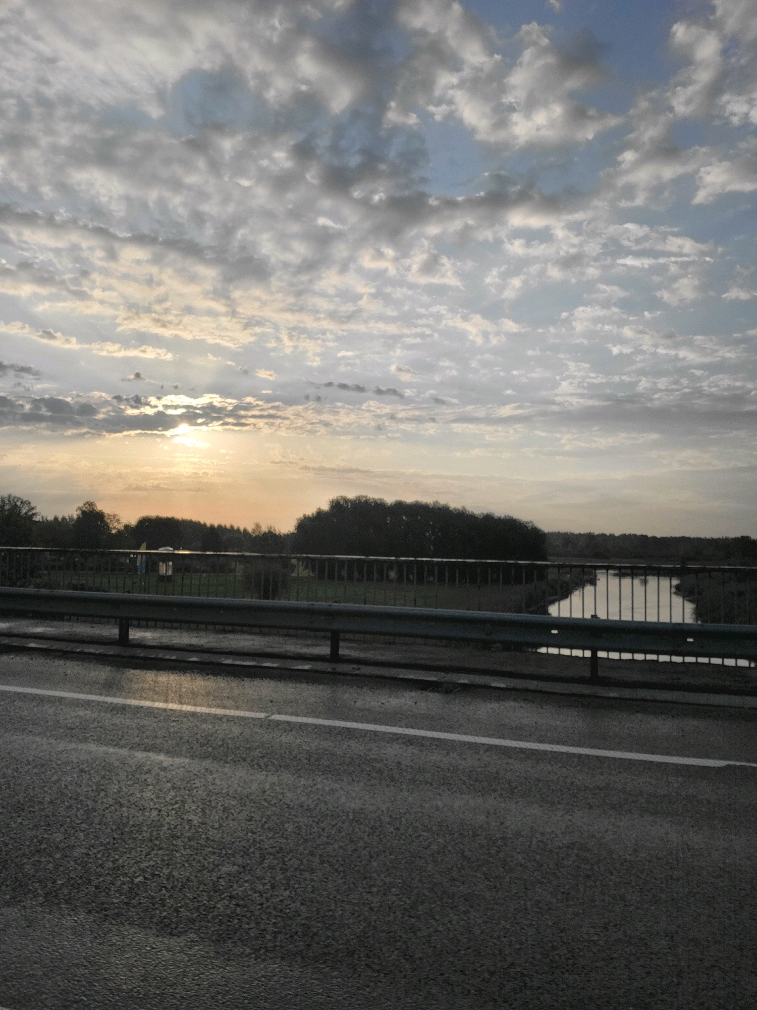 The image depicts a twilight scene over a body of water, likely a river, with a road in the foreground. The sky is filled with scattered clouds, some dark and dramatic, others lighter, with the sun peeking through the clouds on the left side, casting a warm glow. A low-lying treeline runs along the far bank of the river, with a few structures possibly visible amongst the trees. A bridge railing is visible in the middle ground, separating the road from the river view. The road surface in the foreground appears wet, suggesting recent rain or damp conditions. The scene is tranquil and natural, with a focus on the interplay of light and clouds over the water and landscape. The location is specified as Jelgava, Latvia, but no specific landmarks are identifiable from the image alone to confirm this without external information. There are no people or animals visible in the frame, making it a landscape-focused photograph.