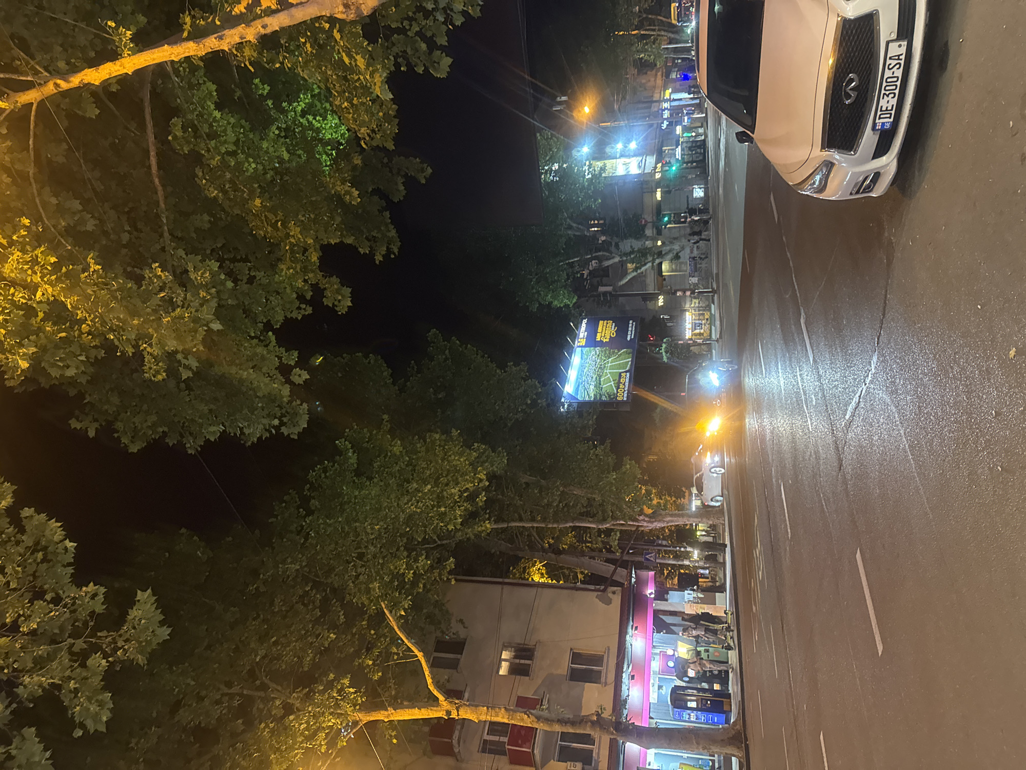 The image captures a street scene in Tbilisi, Georgia, at night. A white Infiniti sedan is parked on the right side of the frame, facing left. Its license plate reads "DE-300-SA". The street is wet, indicating recent rain. Across the street, there is a building with illuminated windows and a storefront featuring bright pink and magenta accents. Several people are visible near the storefront, some standing and others appearing to interact with machines or each other. A street light casts a warm glow on the trees and the building. Further down the street, the headlights of approaching vehicles are visible. Large billboards are present on the buildings, one of which shows an image of a sports field. Trees line the sidewalk, their leaves illuminated by streetlights. The overall atmosphere is that of a typical urban night, with activity and artificial lighting.