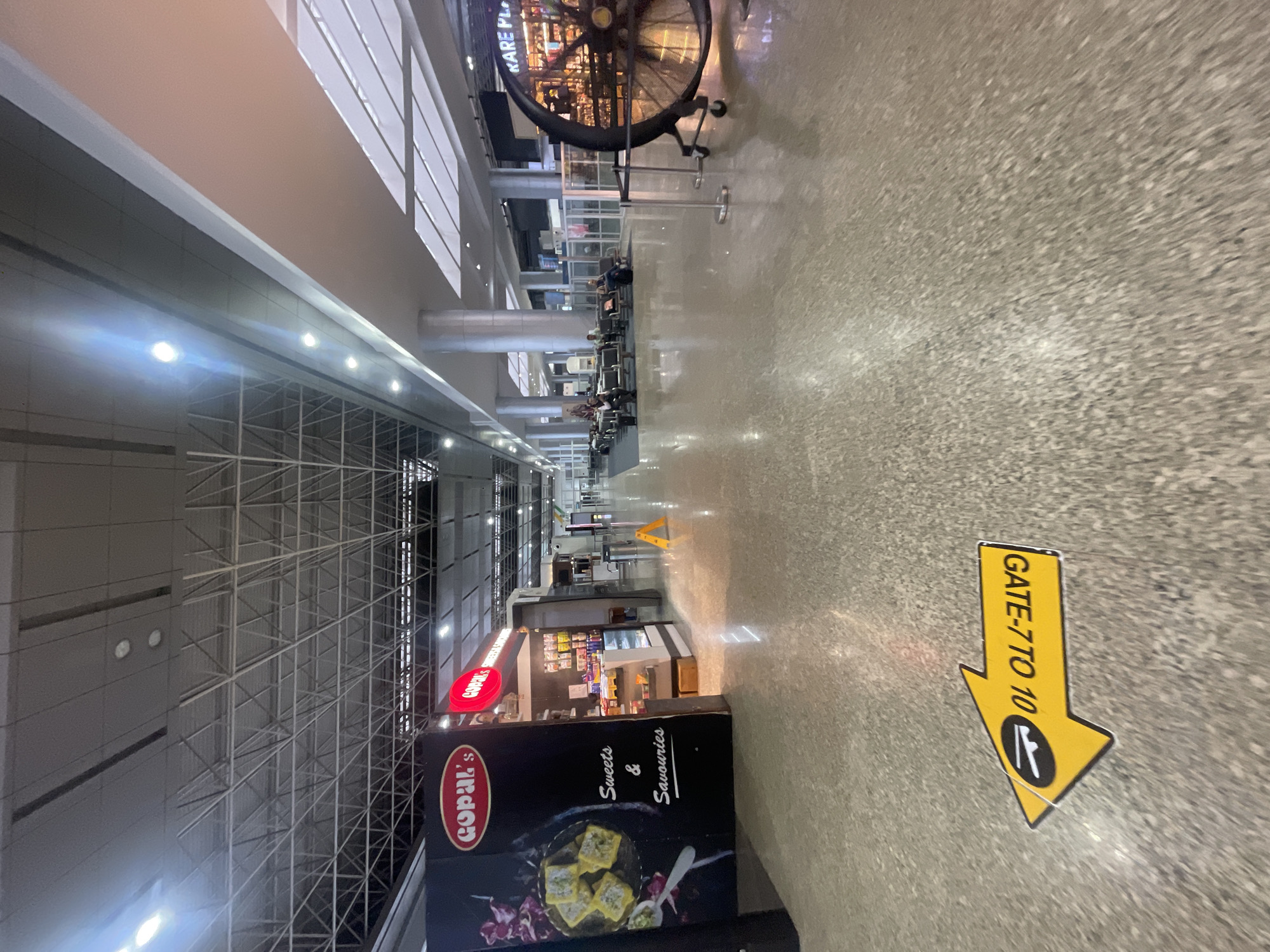This image captures an interior view of the Swami Vivekananda Airport in Raipur Khurd, India. The scene depicts a well-lit airport concourse with a polished floor that reflects the overhead lights and architectural elements. In the foreground, a yellow directional arrow sticker on the floor indicates "GATE 770 10". To the left, a kiosk labeled "GOPAL's Sweets & Savouries" is visible, featuring branding and images of food items. Further into the concourse, rows of seating are arranged for passengers, with several individuals seated and a few standing. Various shops and structures line the concourse in the background, including a large decorative fan-like structure on the right with the text "KARE PL" visible on it. The ceiling features a complex industrial design with exposed metal beams and lighting fixtures. The overall environment suggests a functional and organized airport terminal.