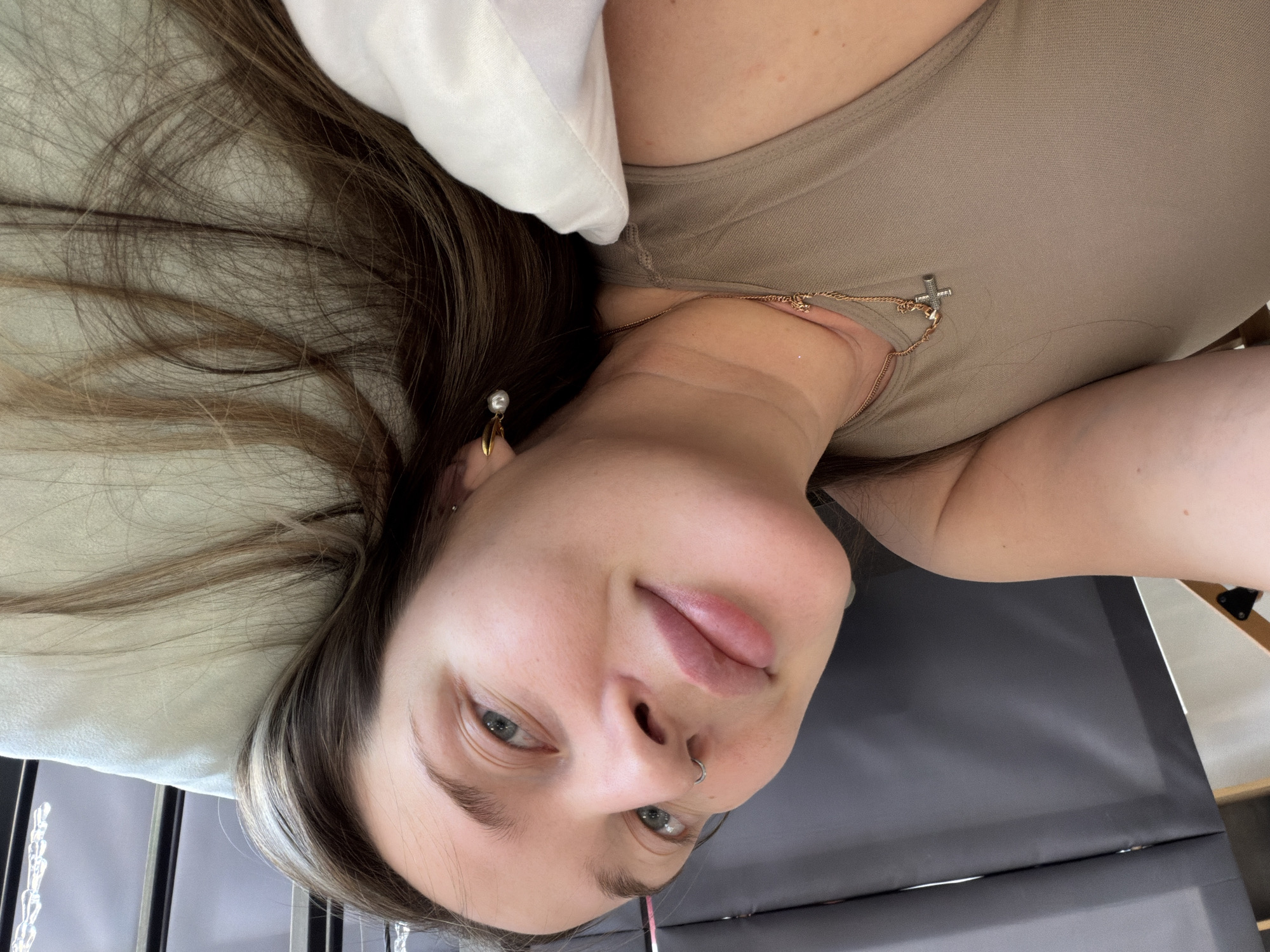 A young woman with light skin and light brown hair with a blonde streak is lying on a grey surface, likely a bed or massage table. She is wearing a tan-colored top and a necklace with a cross pendant. A white pillow is behind her head. She has a nose ring and an earring with a pearl on her left ear. The image appears to be a selfie taken from an overhead perspective. The location, Chaikovsky, Russia, cannot be verified based on the image.