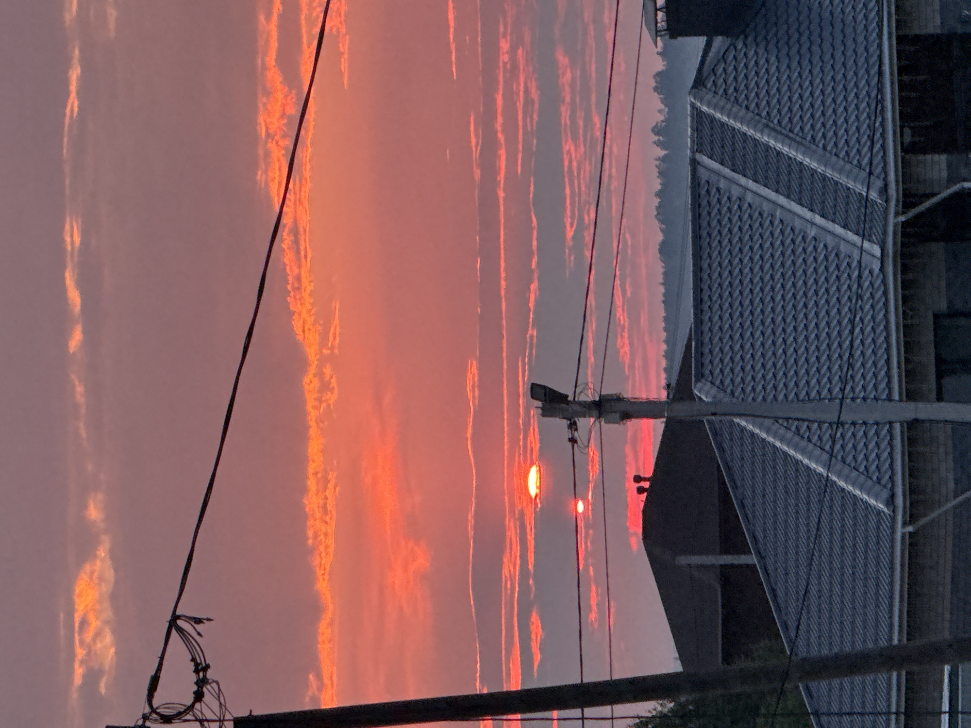 The image depicts a sunset in Izhevsk, Russia, with a vibrant pink and orange sky filled with scattered clouds. The foreground is dominated by the dark silhouettes of rooftops, possibly belonging to houses or buildings, with some appearing to be under construction or in a state of disrepair. Power lines and a utility pole are prominent in the middle ground, stretching across the frame and partially obscuring the view of the sky and the distant treeline. Two suns are visible in the sky, one larger and higher than the other, both appearing as glowing red discs. The lower sun casts a red reflection onto the clouds below it. The overall scene suggests a rural or suburban setting at dusk.