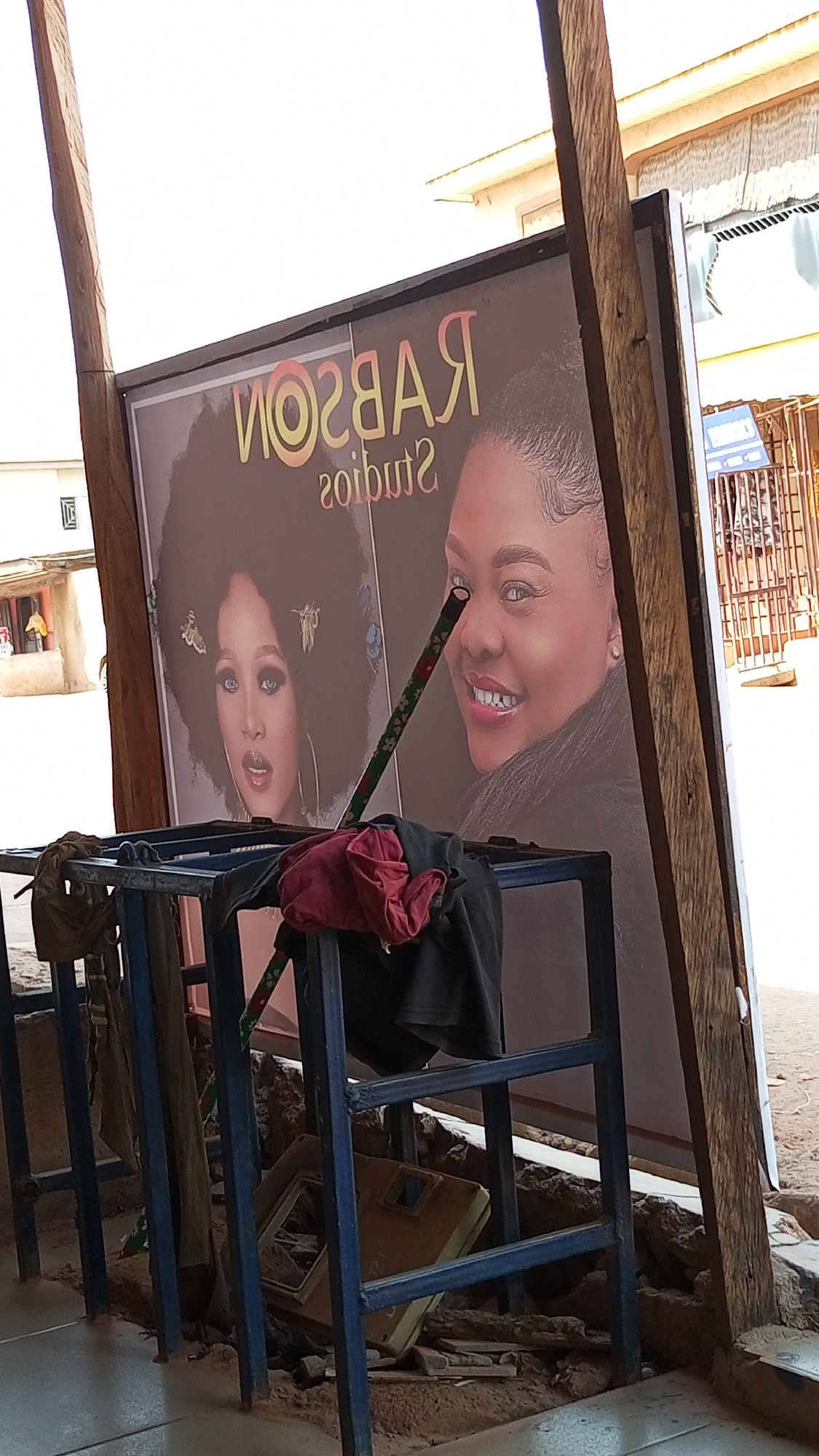 The image shows an outdoor advertisement for "Rabson Studios" in an unidentified city. The advertisement features portraits of two women: one with a large afro hairstyle and the other with a shorter, styled haircut. The sign is propped up by wooden posts and obscured by a blue metal rack with clothing and a cleaning mop leaning against it. Debris is scattered on the ground in front of the sign. In the background, there are buildings and a partially visible street.