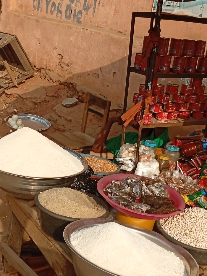 A roadside market displays various food items. Large bowls contain rice, flour, and beans. A red tray holds dried fish. Peanuts are in a nearby dish. Shelves behind the counter are stocked with rows of red canned goods. Jars and small packages of goods are also visible. The setting appears to be a weathered outdoor area, with a worn wall visible in the background.