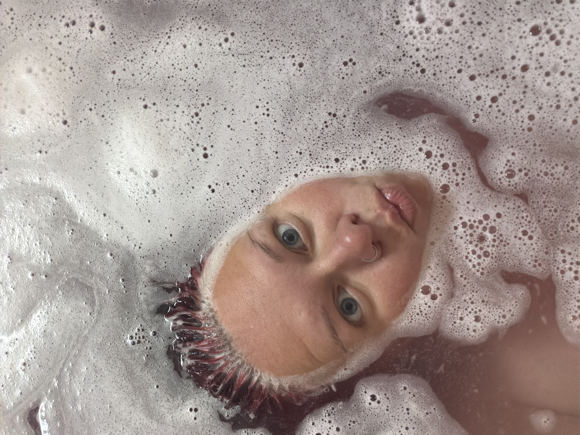 A close-up, overhead shot shows a person's face partially submerged in a bathtub filled with frothy, pinkish-brown water. The individual has light skin, blue eyes, and bright red streaks in their dark hair. A silver hoop is visible in their nose. Their lips are pursed. The water is covered in a thick layer of small bubbles, with some larger bubbles also present. The image captures a moment of personal relaxation or hygiene. The city of Chaikovsky, Russia is not depicted in this image.