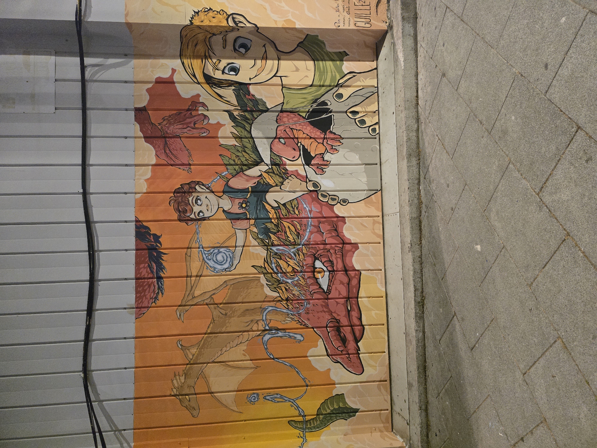 A mural is depicted on a corrugated metal wall adjacent to a paved walkway. The mural features cartoon-style illustrations, including two figures with humanoid features and two dragons. The upper figure has blonde hair and is wearing a green garment. Below, a smaller figure with brown hair holds a swirl of blue energy. A large, red dragon is prominently featured with detailed scales and a single visible eye. A second, smaller dragon is partially visible. The mural uses shades of orange, red, and yellow. To the left of the mural, a black electrical cable runs vertically down the metal wall. The paved walkway consists of gray rectangular stones. The mural is located in Cornellà de Llobregat, Spain.