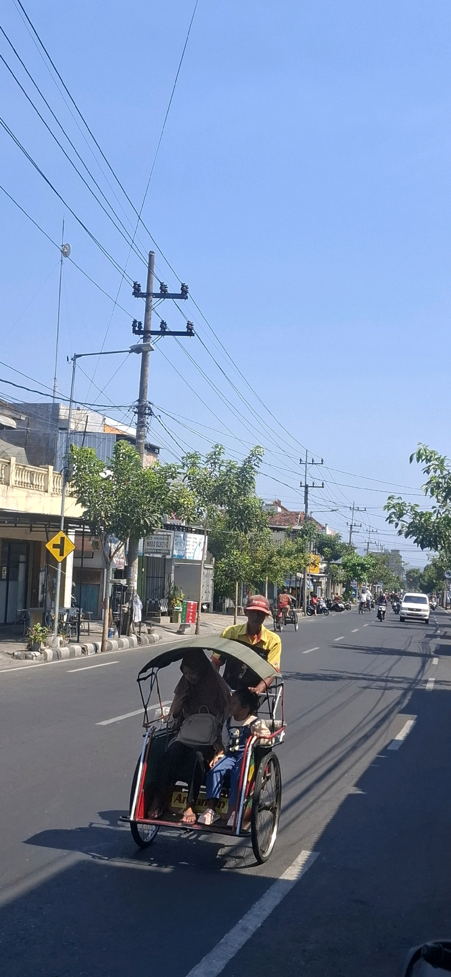 A street scene in Blitar, Indonesia, shows a pedicab in the foreground carrying two passengers, a woman in a hijab and a young girl. The pedicab is being driven by a man wearing a yellow shirt and a red cap. In the background, there are shops and buildings lining the street, with trees and electrical wires overhead. Several other vehicles, including motorcycles and a white van, are visible further down the road, suggesting moderate traffic. The sky is clear and blue, indicating a sunny day. The location appears to be a typical urban street in Blitar, with a mix of commercial establishments and residential buildings.