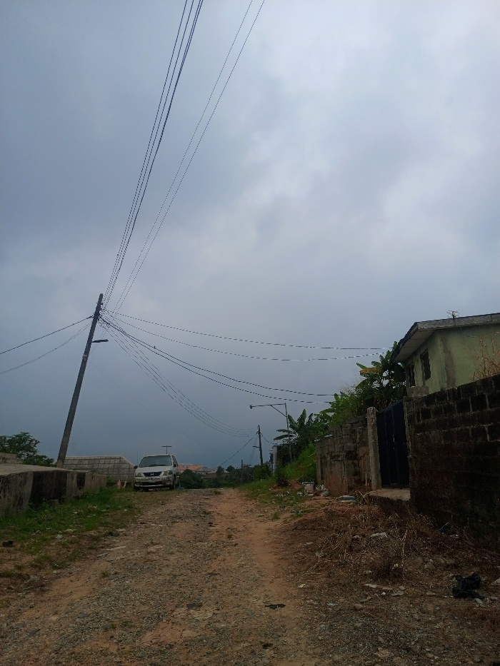 The scene depicts an unpaved, gravel road under an overcast sky in a residential area of Magboro, Nigeria. The sky is uniformly grey, indicating heavy cloud cover. The reddish-brown dirt and gravel road, showing tire tracks, extends into the background.
On the left side, a concrete utility pole stands prominently, supporting multiple overhead power lines that traverse the upper portion of the frame. Below and slightly beyond the pole, a light-colored minivan or SUV is parked facing away from the viewer, situated near a low concrete structure.
On the right side, an unrendered concrete block wall runs parallel to the road. Behind this wall, a light green, single-story building with a dark roof and a roof-mounted antenna is partially visible. A dark blue metal gate is integrated into the wall. Dense tropical foliage, including broad-leaved plants, grows behind the wall and around the building. Further down the road on the right, another utility pole with a street lamp fixture is visible.
No individuals are present. No specific actions or interactions are depicted. The environment consists of sparse dry vegetation and some debris alongside the road.
