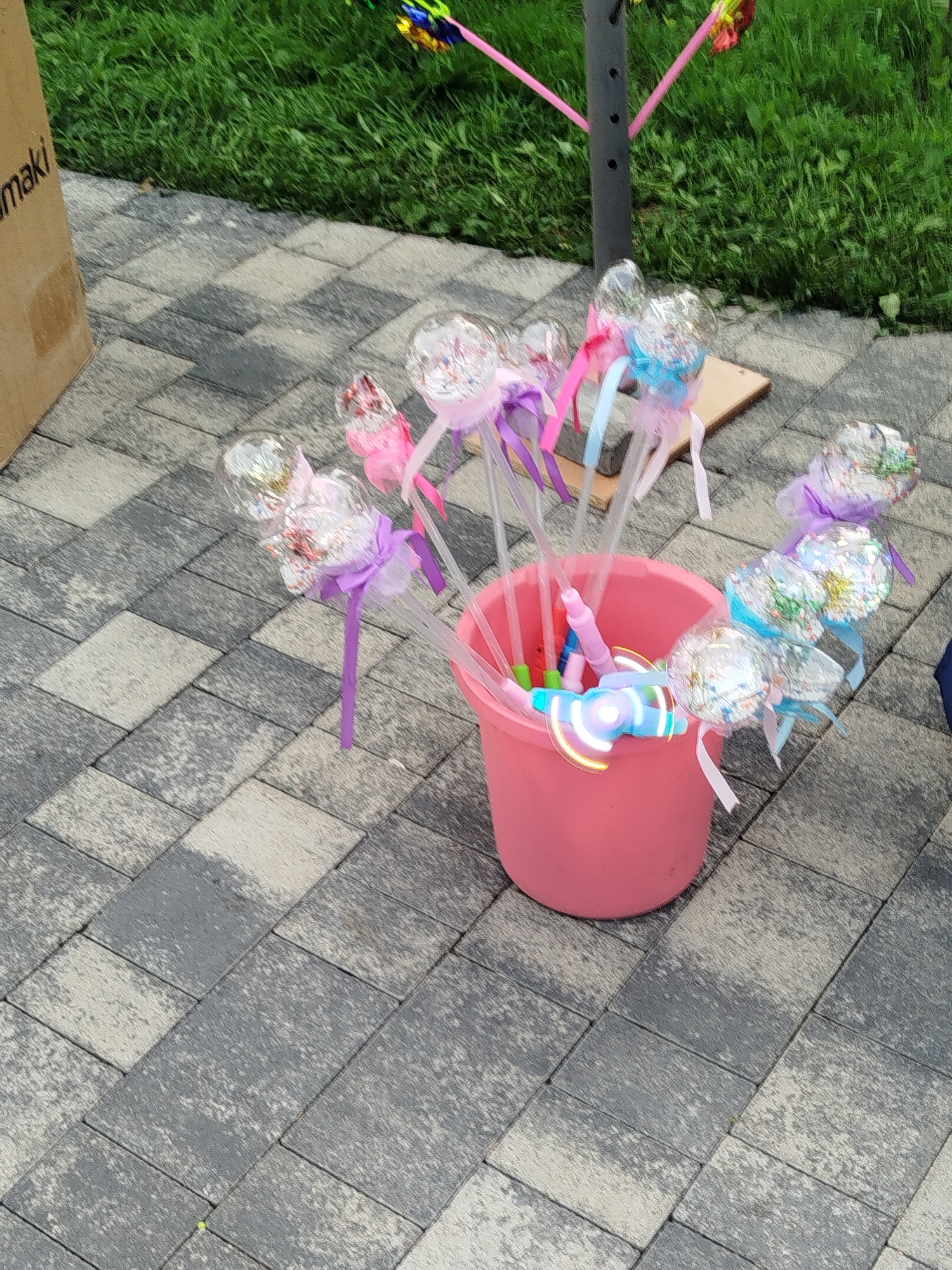 A pink bucket filled with novelty wands sits on a paved surface next to a grassy area. The wands have clear plastic tops filled with glitter and colorful ribbons. One wand in the bucket is lit with blue and pink lights and has a propeller-like attachment. A cardboard box with text that appears to be "maki" is visible to the left of the bucket. In the background, green grass and a black pole with pink streamers suggest an outdoor setting, possibly a park or a street. The paved surface is made of rectangular blocks in varying shades of gray and beige. No people are visible in the frame. The scene is devoid of any specific visual cues that would pinpoint its location to Zavodoukovsk, Russia.