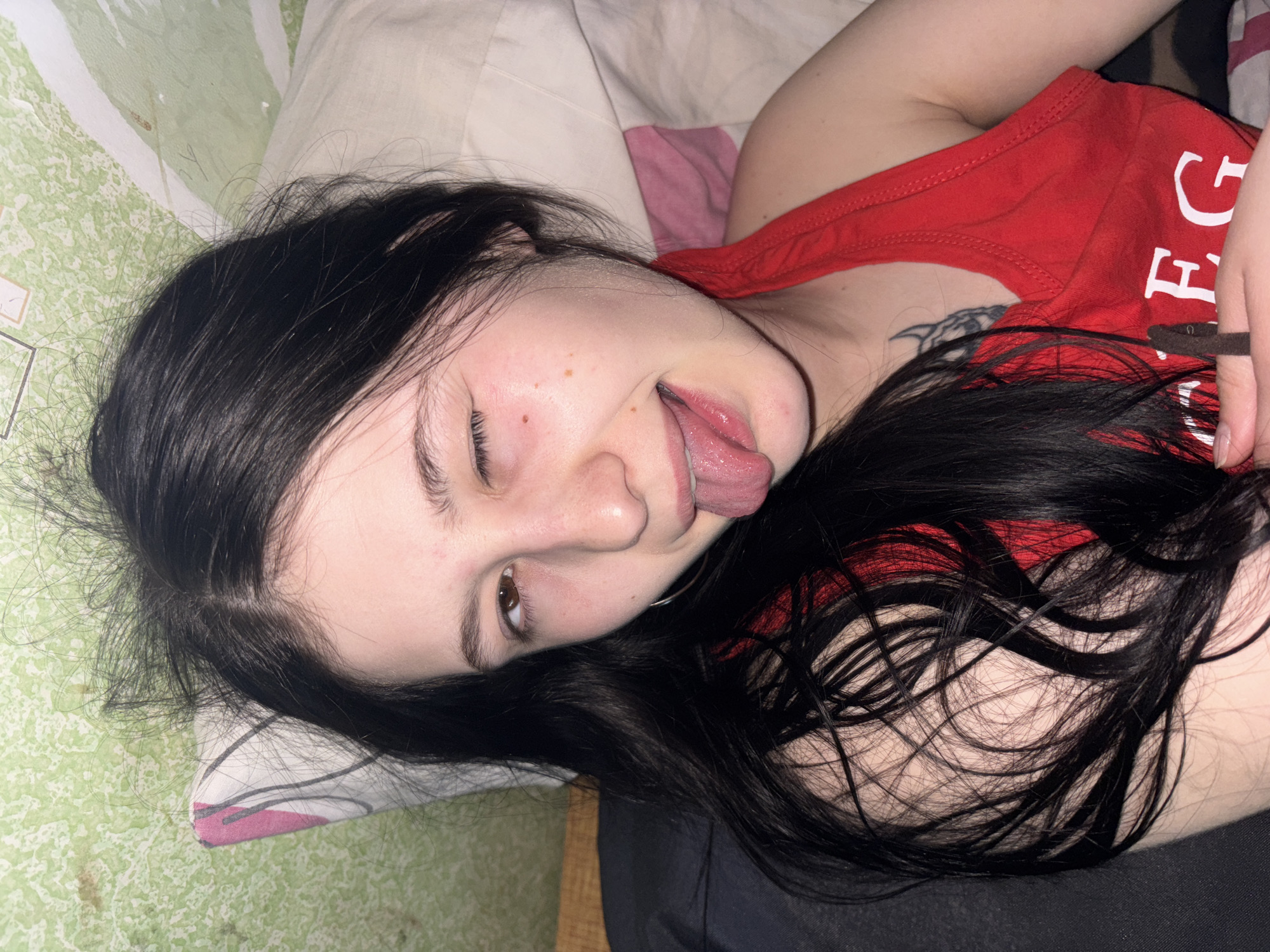 The image displays a young female subject reclining indoors, located in the city of Фоки, Russia. She has long, dark hair, light skin, and is wearing a red tank top with partially visible white lettering ("EG"). A dark, abstract tattoo is visible on her left shoulder area. Her right eye is closed in a wink, and her tongue is extended from her mouth. Her left eye is open, revealing a brown iris, and a silver hoop earring is present in her left ear. Small reddish marks are visible on her right cheek. She is positioned against a white pillow with pink and dark patterns, which rests against a light green wall featuring textured wallpaper. A dark fabric element is present to the lower left of the frame.