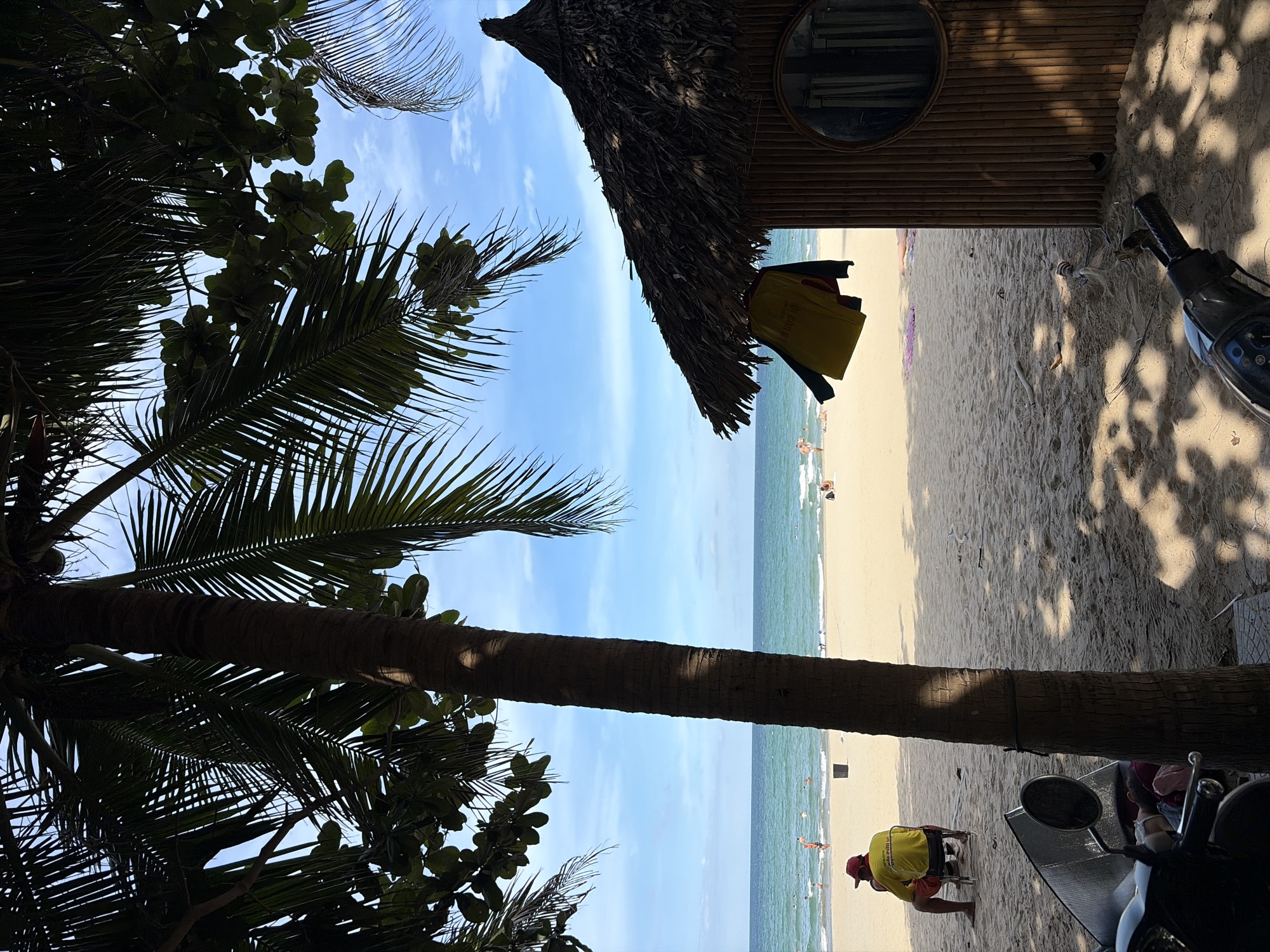 This image depicts a beach scene in Da Nang, Vietnam. The foreground features a palm tree trunk and a motorcycle. In the mid-ground, a thatched-roof hut with a circular mirror is visible on the right. A person wearing a red hat and a yellow shirt sits on a chair on the left, facing the ocean. The beach is sandy, with several people scattered across it and in the turquoise water. The sky is blue with some clouds. The overall ambiance suggests a sunny day at a coastal resort.