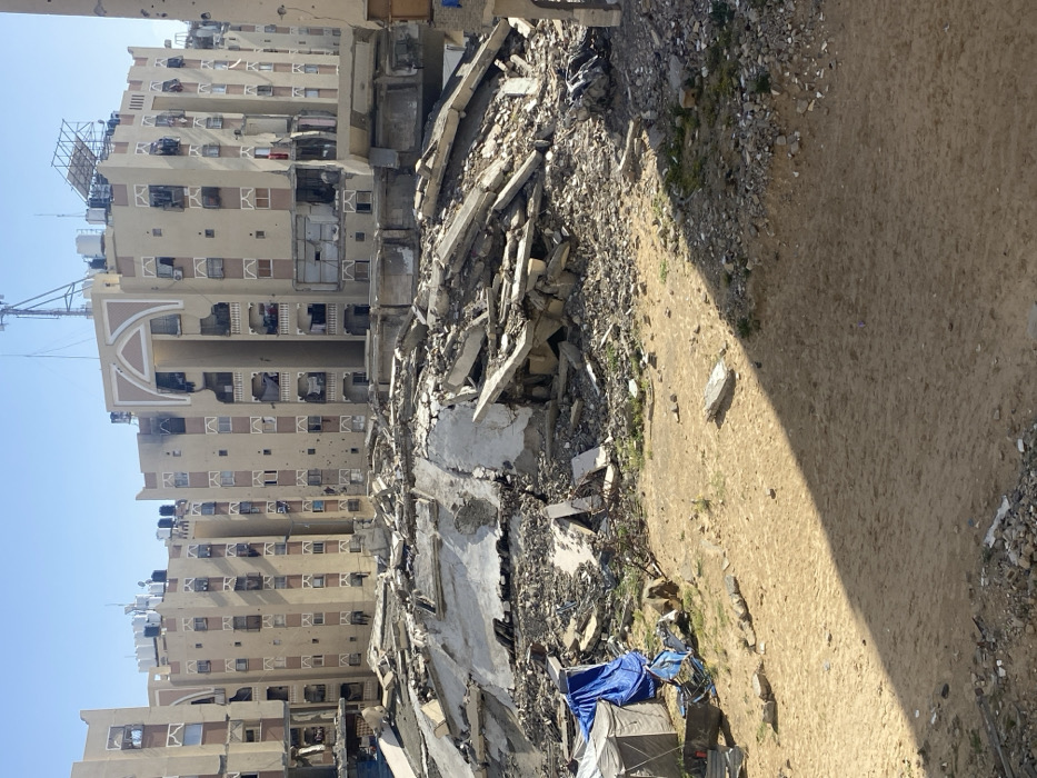 The image displays a scene of extensive urban destruction in Khan Yunis, Palestinian Territory, under a clear blue sky. In the foreground, a light brown, sandy ground is scattered with small rocks and minimal sparse vegetation, bisected by a sharp diagonal shadow cast from the left. To the far left, a grey tent and a structure covered by a blue tarp are visible amidst debris. The midground is dominated by a large, irregular mound of concrete rubble, twisted rebar, and other fragmented building materials, indicative of a completely collapsed structure. This debris pile rises significantly, partially obscuring the lower levels of the background buildings. In the background, a row of multi-story residential buildings, constructed with light beige facades and darker brown accents, exhibits widespread severe damage. These buildings show numerous shattered or missing windows, damaged balconies, and extensive pockmarks on their facades from impacts. Some areas feature black scorch marks, suggesting fire damage. Multiple black water tanks and communication antennas are visible on the rooftops. No people are present, and no actions or interactions are depicted within the scene.
