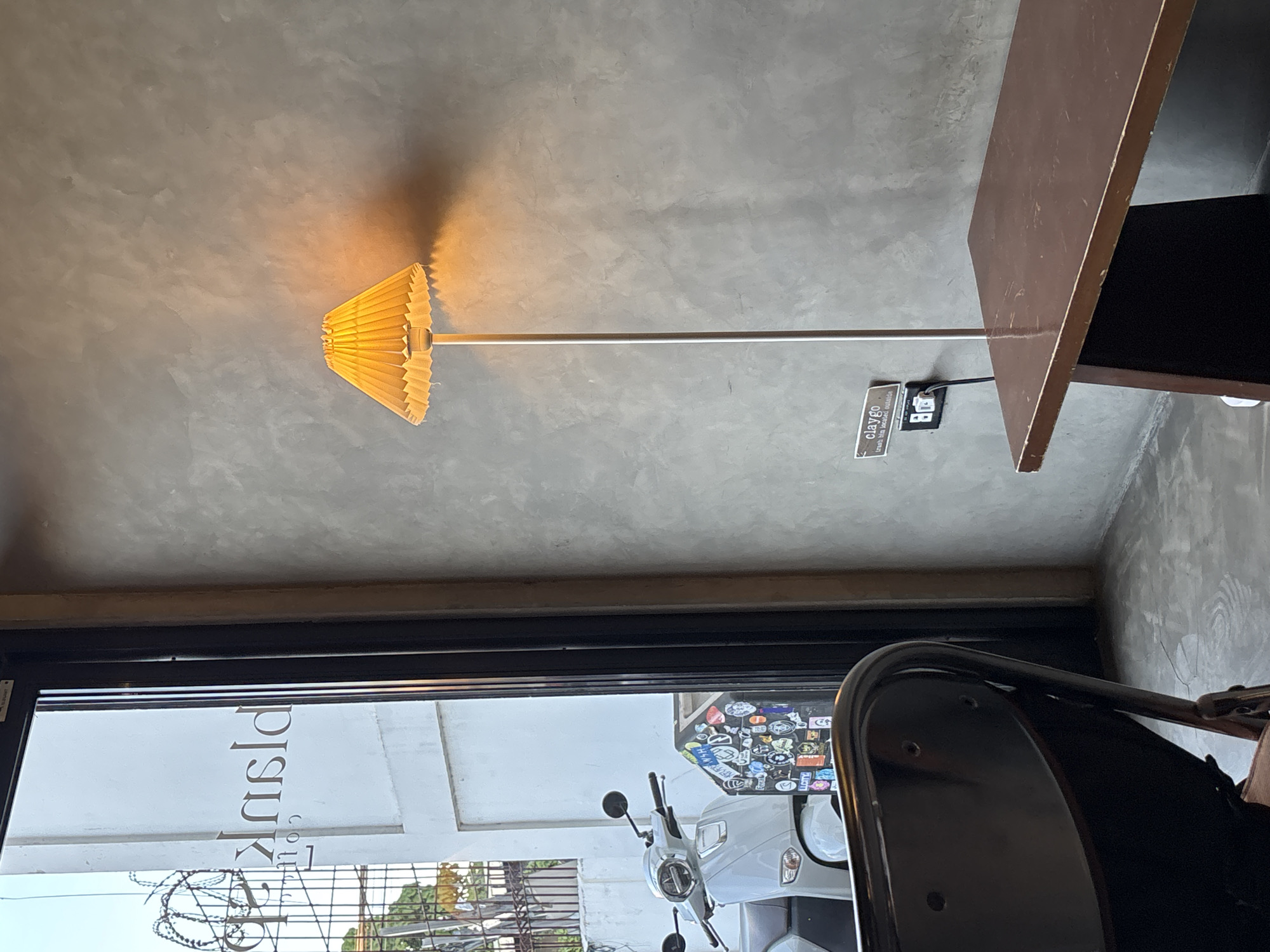 A floor lamp with a pleated yellow lampshade illuminates a textured concrete wall in this indoor scene, casting a warm glow and a distinct shadow. The lamp's slender silver pole extends from the floor, with a small black electrical outlet mounted on the wall near its base. A small tag attached to the outlet reads "claygo" and below it, "trash bin located outside." To the right of the lamp, the edge of a dark brown wooden table is visible, with a dark surface underneath it.
Through a large glass window or door in the foreground, a glimpse of the outside world is provided. The window has a black frame. Reflected on the glass is a partial view of a white building with stylized text that appears to read "collections" in a vertical orientation, with other lettering above and below. A portion of a light-colored scooter, adorned with numerous stickers, is parked outside. A metal grate or fence is also visible in the outdoor scene. The overall impression is of a contemporary, perhaps minimalist, interior with a view into an urban setting. The lighting suggests it could be daytime, possibly late afternoon or early evening given the warm lamp light.