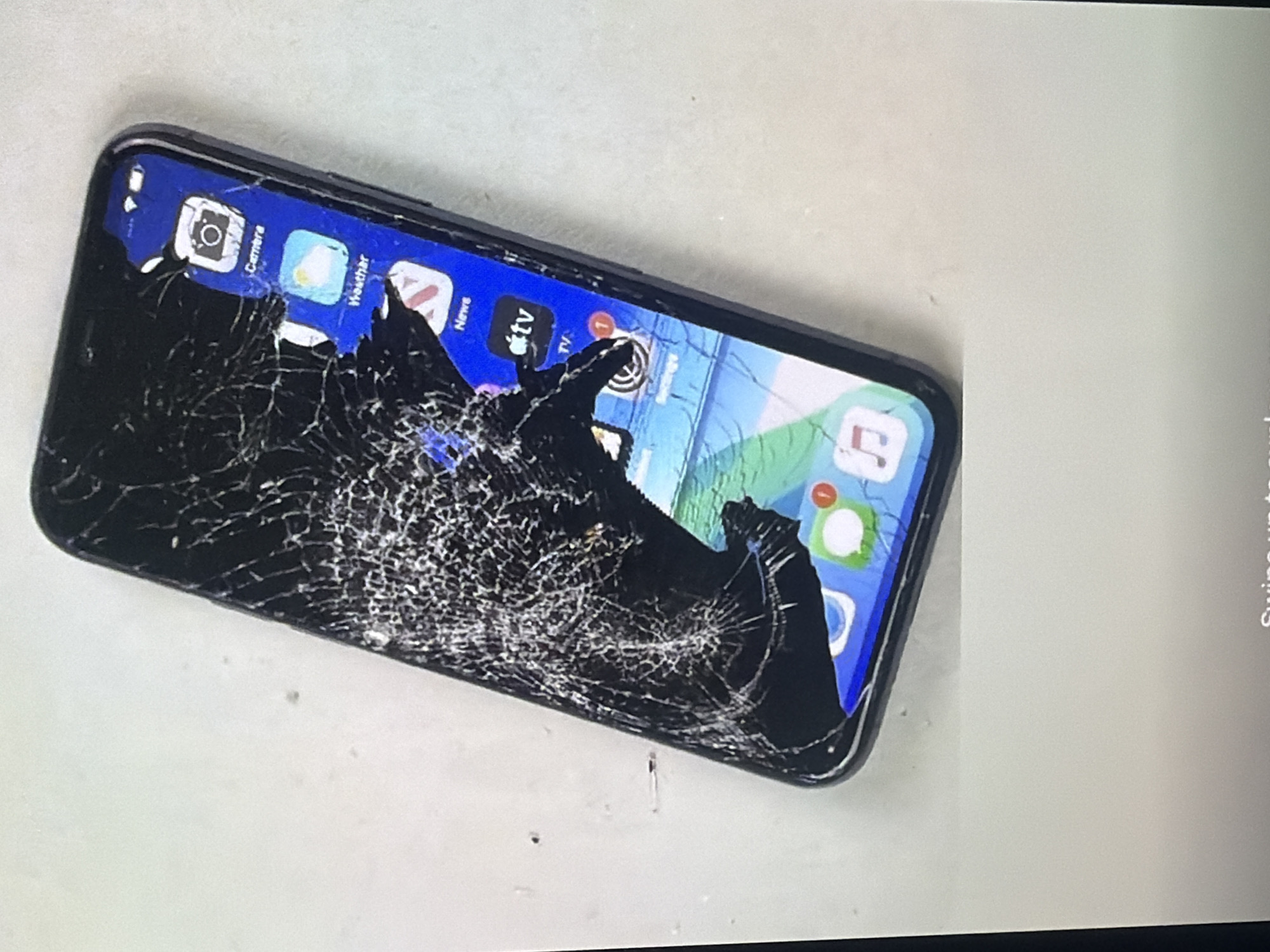 The image displays a severely damaged smartphone lying on a light-colored, textured surface. The screen is heavily cracked, with large sections of the glass missing, revealing the underlying display and internal components. Several app icons are partially visible on the screen, including "Camera," "Weather," "News," "TV," "Settings," and "Messages." The phone appears to be a dark color, possibly black or dark gray, with a metallic frame. The background is plain and out of focus, suggesting a neutral setting. There are no people visible in the image, and no specific geographical markers are evident to indicate a location in Bukuru, Nigeria.