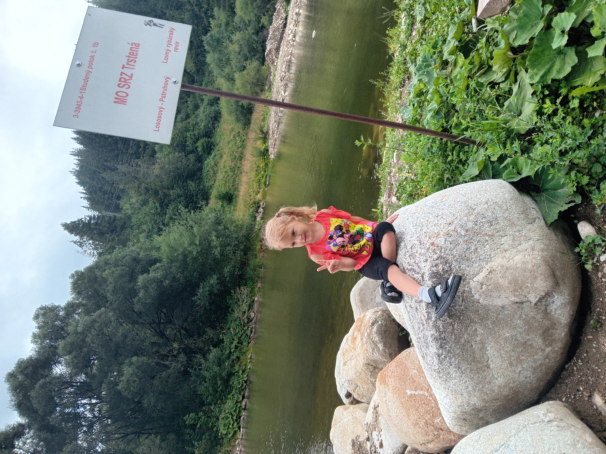 A young Caucasian girl with blonde pigtails, wearing a pink t-shirt with cartoon characters and black shorts, is sitting on a large boulder by the Orava River. She is making a peace sign with her right hand. The river is green and calm, reflecting the cloudy sky. In the background, dense green forest covers the hills. A white sign with Slovak text and a small illustration of a fisherman is visible to the right of the girl. The location is identified as the Orava River near Habovka, Slovakia, a popular fishing area.