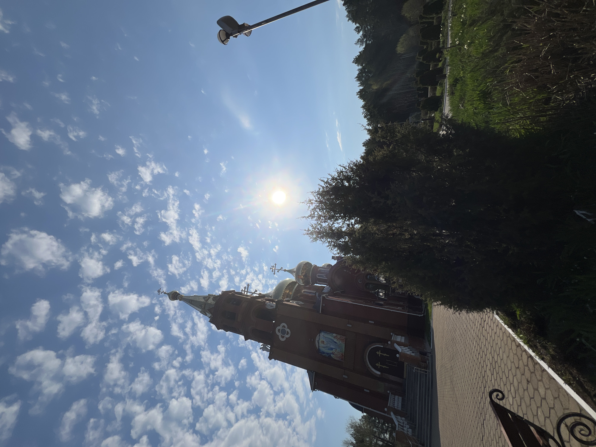 The image is taken in Chaikovsky, Russia, and features the Holy Temple of the Presentation of the Lord Church. The church is constructed of red brick with golden domes and towers reaching into a bright blue sky dotted with white clouds. The sun is positioned high in the sky, casting a glare. To the right of the church, there is a pathway made of patterned paving stones, bordered by greenery including trees and trimmed shrubs. A lamppost is also visible in the upper left corner.