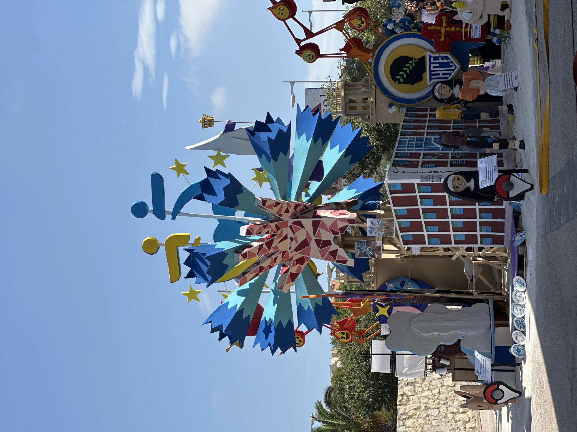 The image depicts an outdoor display of colorful, elaborate structures, likely part of a festival or exhibition. The central and largest structure is a large, multi-colored, abstract design resembling a starburst or a fan, composed of geometric shapes in shades of blue, red, and yellow, adorned with yellow stars. Flanking this central piece are various figures and smaller structures. To the left, a grey, cloud-like sculpture stands near a cutout of a character resembling Eevee from Pokemon. To the right, a ferris wheel-like structure with cartoonish faces can be seen. A building facade with red and blue squares is visible behind the central display, and several figures, including what appear to be nuns and children, are positioned in front of it. In the background, trees, buildings, and a clear blue sky with scattered clouds are present. The ground appears to be sandy or dusty.