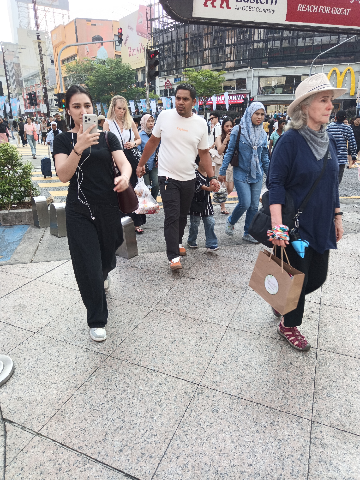 The image depicts a street scene in Kuala Lumpur, Malaysia, likely in a busy commercial area such as Bukit Bintang given the architecture and the presence of commercial signage. Several pedestrians are visible, walking on a tiled sidewalk. In the foreground, a young woman in a black t-shirt and black pants is holding a smartphone in her left hand, wearing earbuds, and carrying a brown shoulder bag. To her right, a man in a white t-shirt and black pants walks with his right arm outstretched, possibly holding hands with someone off-frame. Further to the right, an older woman wearing a beige hat and a dark blue long-sleeved top walks with a shopping bag in her right hand. Other individuals, including a woman in a blue headscarf and denim jacket, are also walking in the same direction. The background features multi-story buildings with prominent commercial advertisements, including a McDonald's sign. Traffic lights and street infrastructure are also visible, indicating an urban setting. The overall atmosphere suggests a bustling day in a city center.