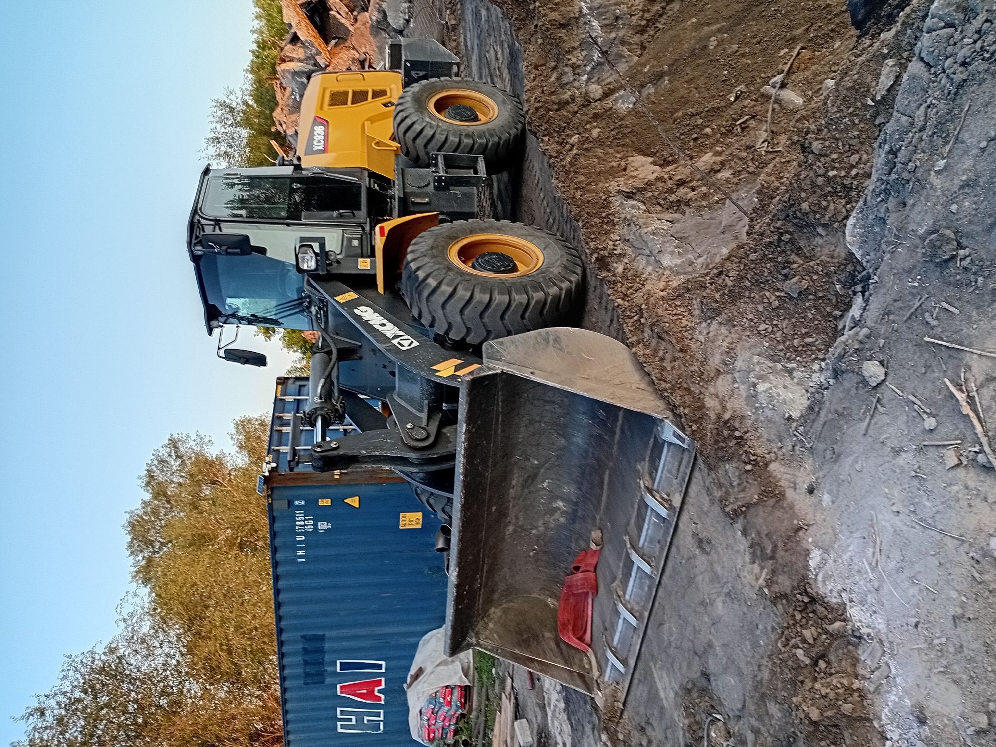 A yellow XCMG XC938 wheel loader is parked on a construction site in Semey, Kazakhstan. The loader has its large bucket lowered to the ground, filled with dirt. In the background, a large blue shipping container with "HAI" written on it is visible, along with trees and a pile of rocks. The sky is clear and blue.