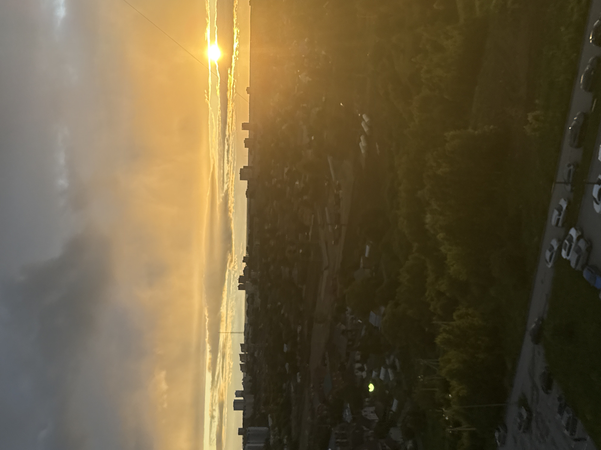 This is a vertically oriented, low-angle, aerial photograph taken from a high vantage point, possibly a building or drone, overlooking a city at sunset. The sun, a bright, intense orb, is positioned on the left side of the frame, casting a warm, golden glow across the sky. Wispy clouds, illuminated by the setting sun, streak across the sky in hues of yellow and white.
Below the sky, a dense urban landscape unfolds. Buildings of varying heights are visible, clustered together and interspersed with areas of lush green trees, suggesting a mix of residential and commercial zones, possibly a park or forested area within the city. The overall impression of the city is one of a sprawling, densely populated area.
On the right side of the image, a road or street is visible, lined with several parked cars. The cars are dark in color and vary in size and shape, indicating a typical urban street scene.
The time of day is clearly sunset, indicated by the brilliant, low-angled sun and the warm, diffused lighting. The weather appears to be clear with some scattered clouds. The location is implied to be a city in Perm, Russia, based on the provided context. There is no visible text in the image. The overall mood conveyed is one of serene beauty and the calm transition from day to night over an urban environment.
