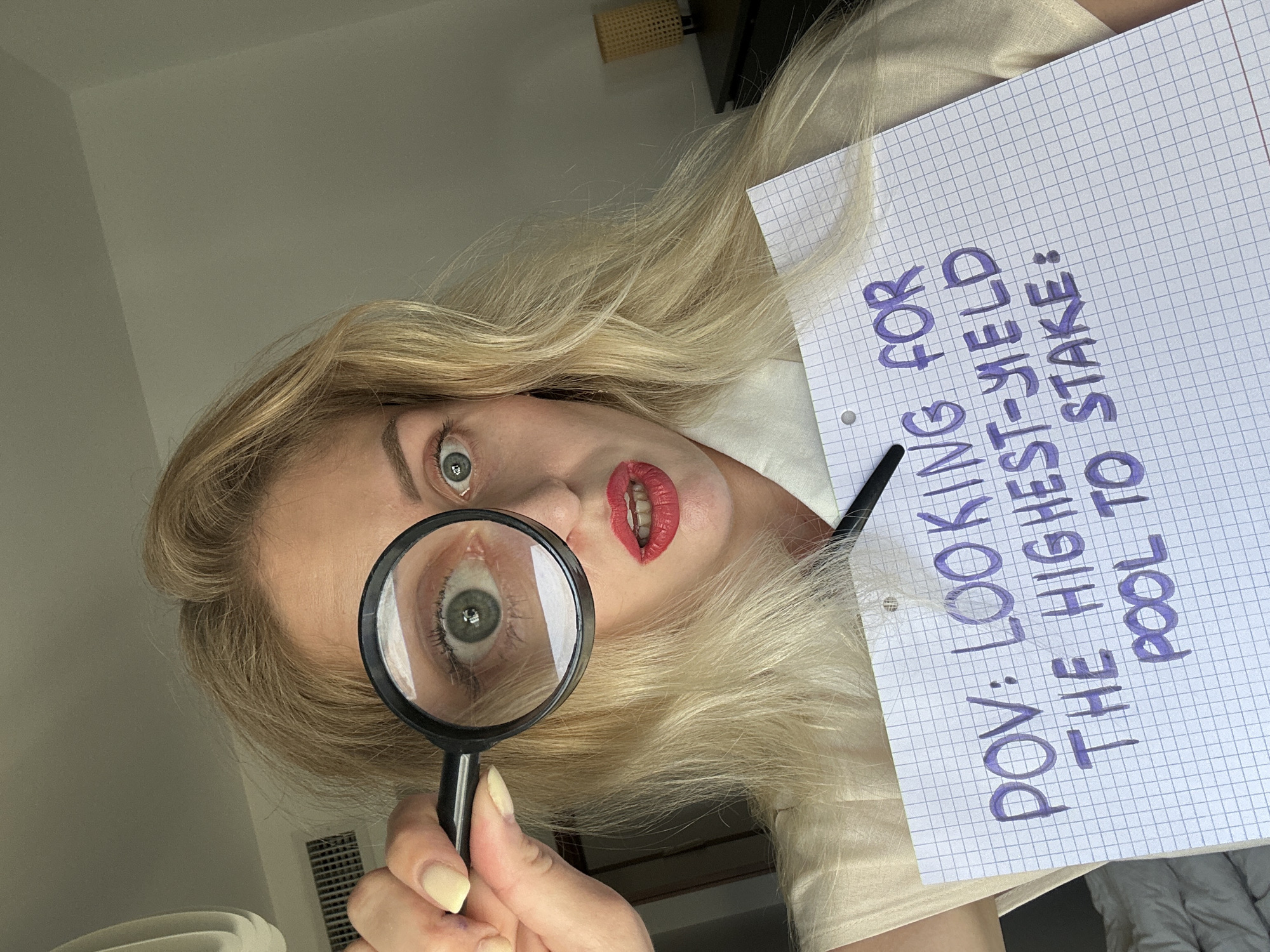 A woman is holding a magnifying glass to her left eye, looking directly at the camera. She has blonde hair and is wearing red lipstick and a white shirt. In her right hand, she is holding a piece of graph paper with the words "POV: LOOKING FOR THE HIGHEST-YIELD POOL TO STAKE:" written on it in purple marker. The background appears to be an indoor setting, possibly a bedroom, with a bed visible on the left and a dresser with a lamp on the right. There is no visible indication of the location being in Petah Tikva, Israel.