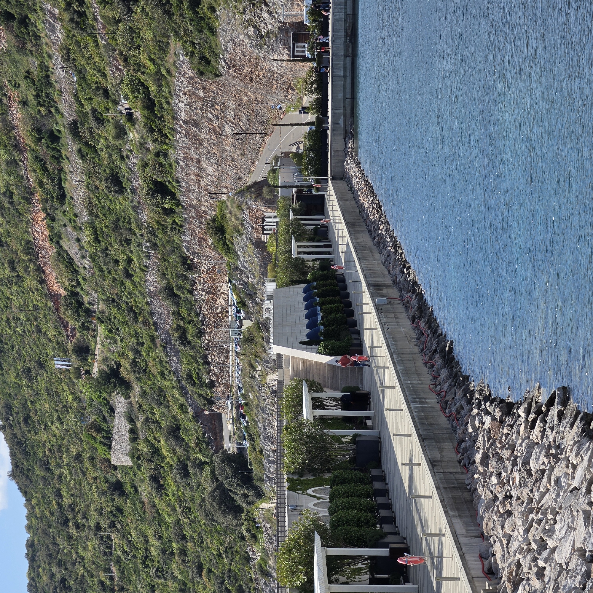 The image presents a picturesque coastal scene in Radovići, Montenegro, showcasing a modern waterfront development at the base of a verdant mountain and alongside a calm body of water. The main subjects include a steeply rising mountain slope, a wide promenade, various resort-style structures, and the clear blue sea.
The mountain, occupying the left side of the frame, is heavily covered with lush green vegetation, interspersed with visible patches of reddish-brown and grey rock faces. A paved road winds along the mountain's lower contours, featuring parked cars and power lines.
The foreground and midground are dominated by a spacious, light-colored promenade, likely made of concrete or stone. This promenade hosts several contemporary buildings with white facades, flat roofs, and open-air sections supported by white columns. These areas are furnished with rows of dark lounge chairs and bright blue umbrellas, suggesting outdoor seating or a pool deck. Manicured green bushes in planters also line the promenade. Access to different levels is provided by wide staircases.
On the far right, the serene, deep blue water of a bay or sea stretches out, reflecting sunlight with numerous small sparkles on its surface. A rugged breakwater, constructed from large, grey rocks, forms the boundary between the promenade and the water. Red lifebuoys on white poles are visible along the promenade's edge, indicating safety provisions.
Several people are engaged in leisure activities on the promenade. One individual in a red shirt and blue shorts is riding a red bicycle, while another person in red stands near a lifebuoy. Other figures are seen walking or standing, enjoying the sunny environment.
The scene is captured during bright daylight, indicated by clear shadows and the sparkling water. The sky, partially visible in the top left, is blue with a few scattered white clouds, suggesting pleasant and clear weather. The overall atmosphere is one of relaxation and recreation. No discernible text is visible in the image.