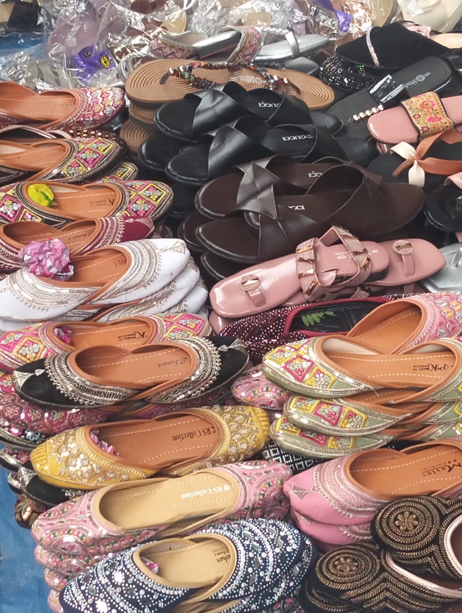 Captured in Dhaka, Bangladesh, this image features a collection of footwear. The assortment includes traditional embroidered Khussa shoes in a variety of colors like pink, yellow, and blue. There are also a variety of modern sandals and slip-on shoes, some bearing brands like Gucci. The arrangement of the shoes appears to be a display for sale, possibly in a market or retail setting. The scene is filled with shoes, emphasizing the abundance and variety of the footwear available.