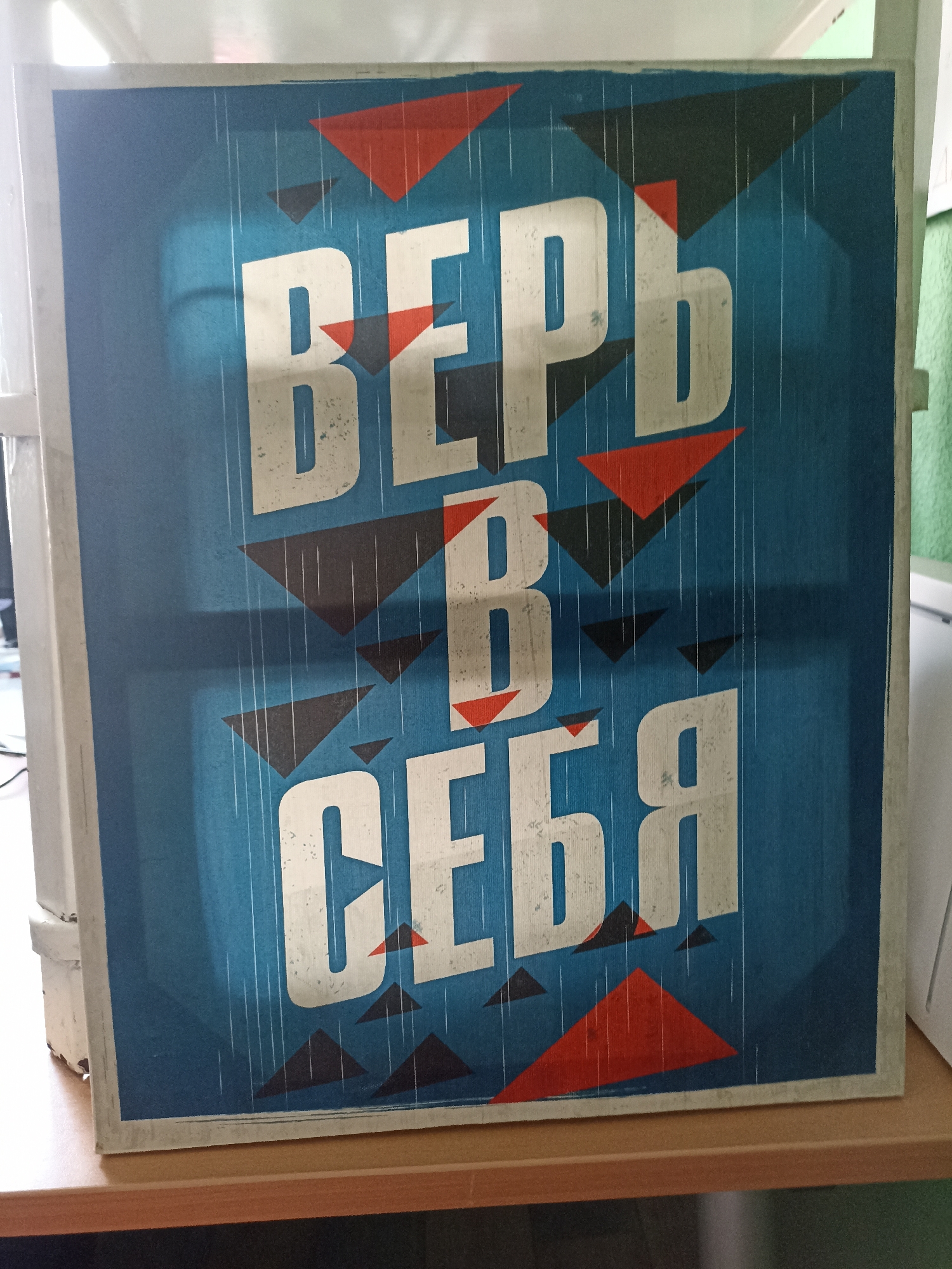 A framed poster with the Russian phrase "Верь в себя" ("Believe in yourself") is displayed on a light-colored shelf. The poster features large, white, distressed lettering against a blue background with stylized red and black triangles and thin white lines resembling rain. The poster is angled slightly upwards, resting on a wooden surface. The surrounding environment is indistinct, with hints of white and green in the background and a white vertical element on the left. The lighting appears to be artificial, casting subtle shadows. The location is identified as Novosibirsk, Russia, but no specific landmarks or identifying features of that city are visible within the frame.