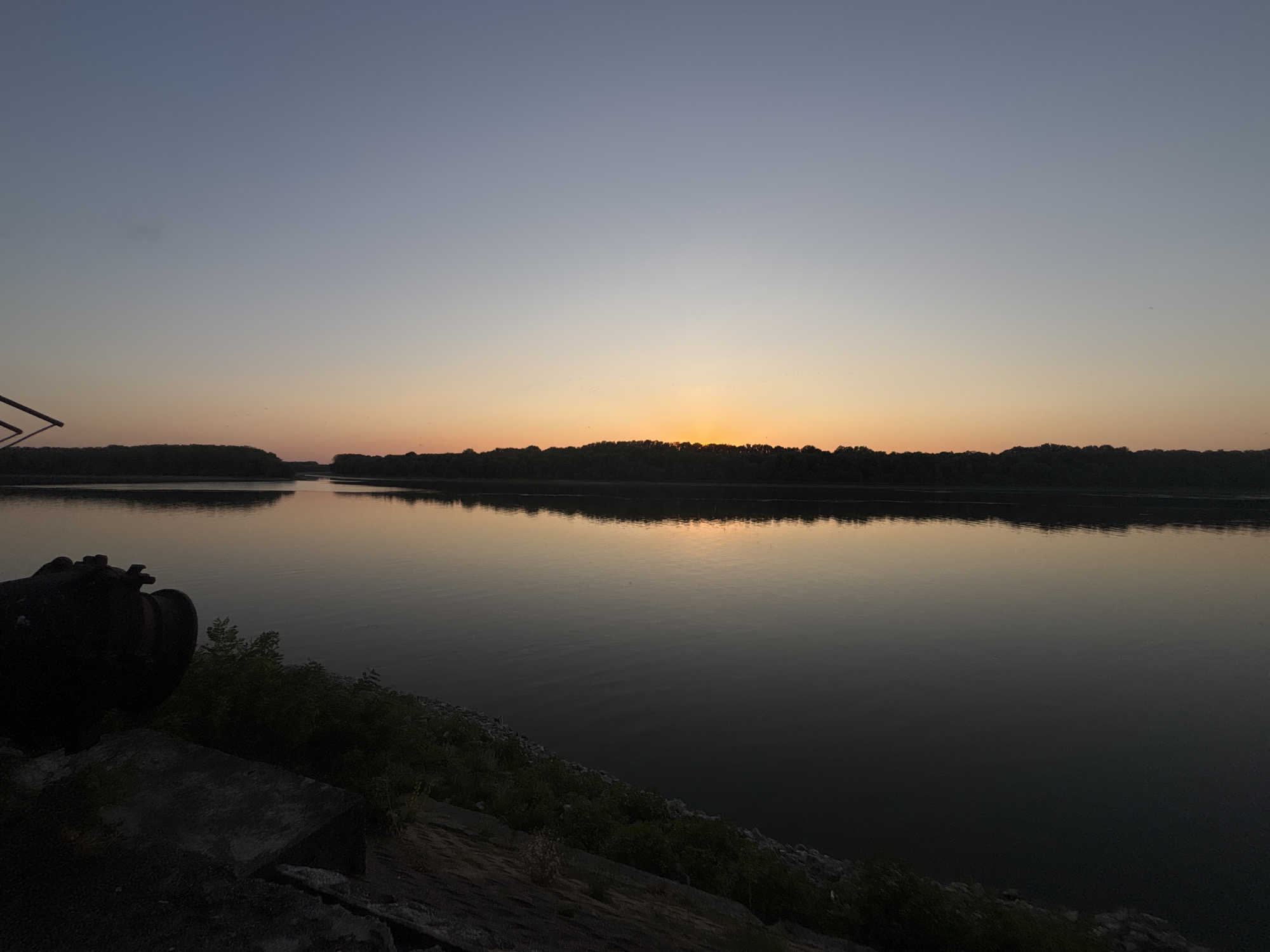 The image depicts a tranquil river scene at dusk. A silhouetted tree line borders the far bank of the wide, dark river, which reflects the soft hues of the sunset. The sky transitions from a pale blue at the top to a warm orange near the horizon, where the sun is just setting. In the foreground, on the left bank, a large, dark, industrial-looking pipe juts out from a concrete structure. Overgrown vegetation, a mix of grass and bushes, surrounds the pipe and concrete. The overall atmosphere is calm and serene, suggestive of an outdoor, natural environment. The location is the river in Marten, Bulgaria.